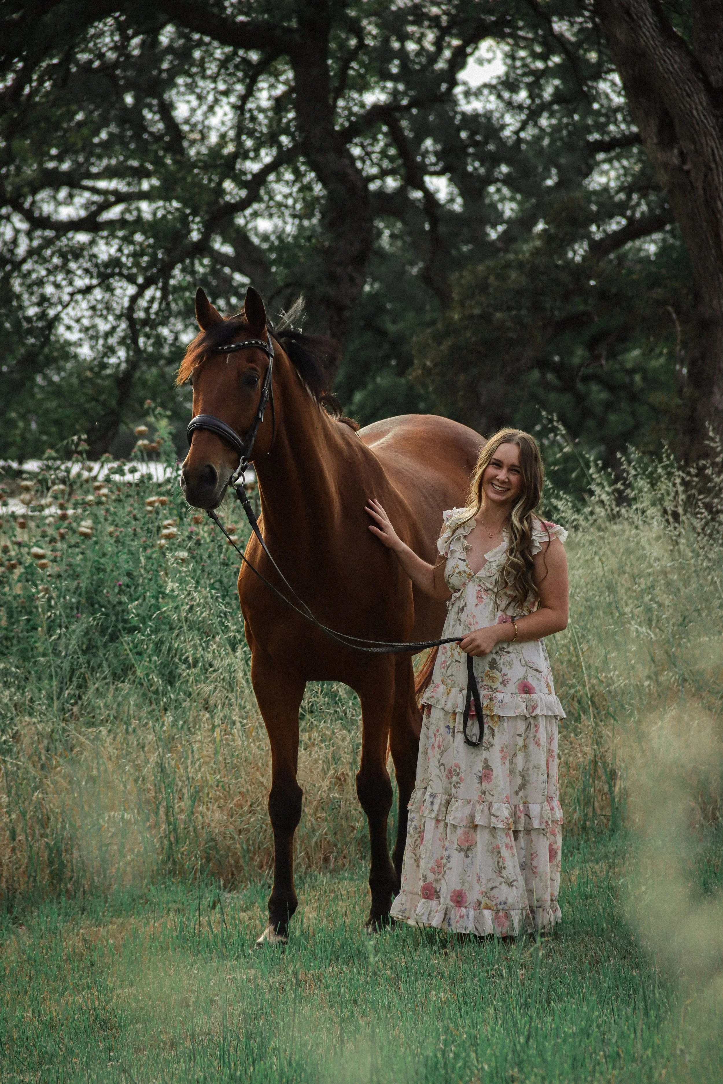A woman with long wavy hair wearing a long floral dress standing next to a brown horse in a grassy field with trees in the background.