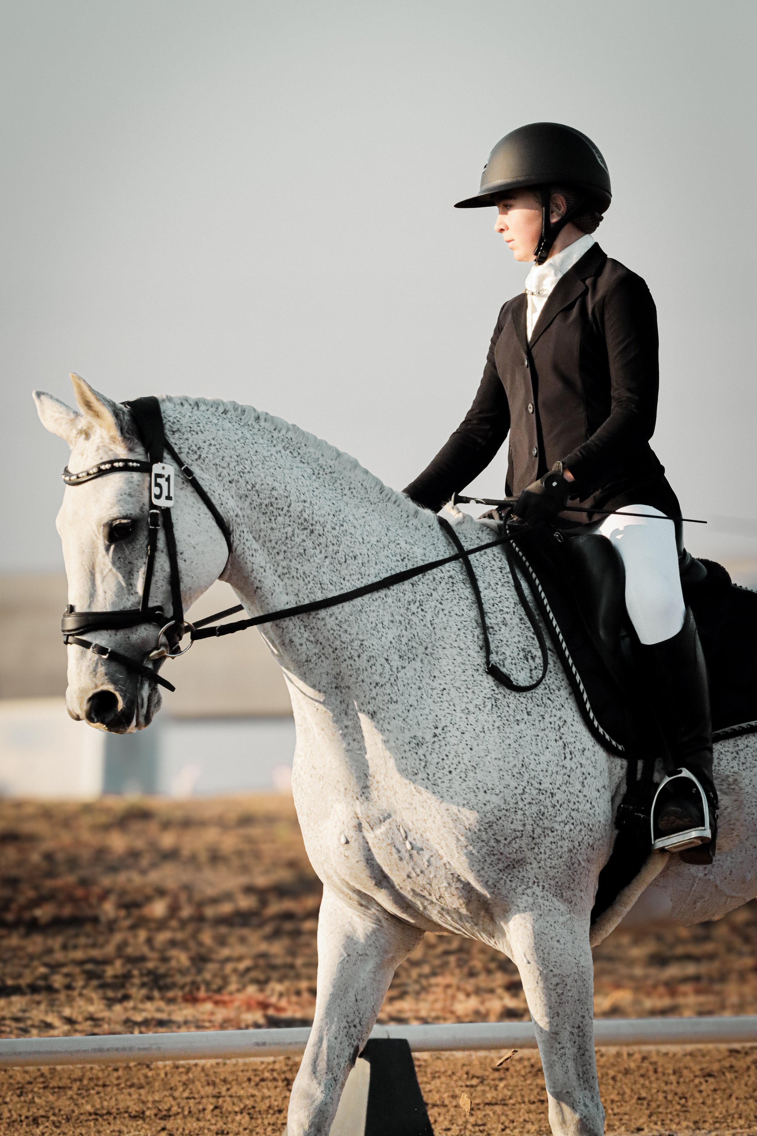 A female equestrian rider wearing a black riding helmet, black jacket, white shirt, and gloves, riding a white horse with speckled gray markings during a show or training session.