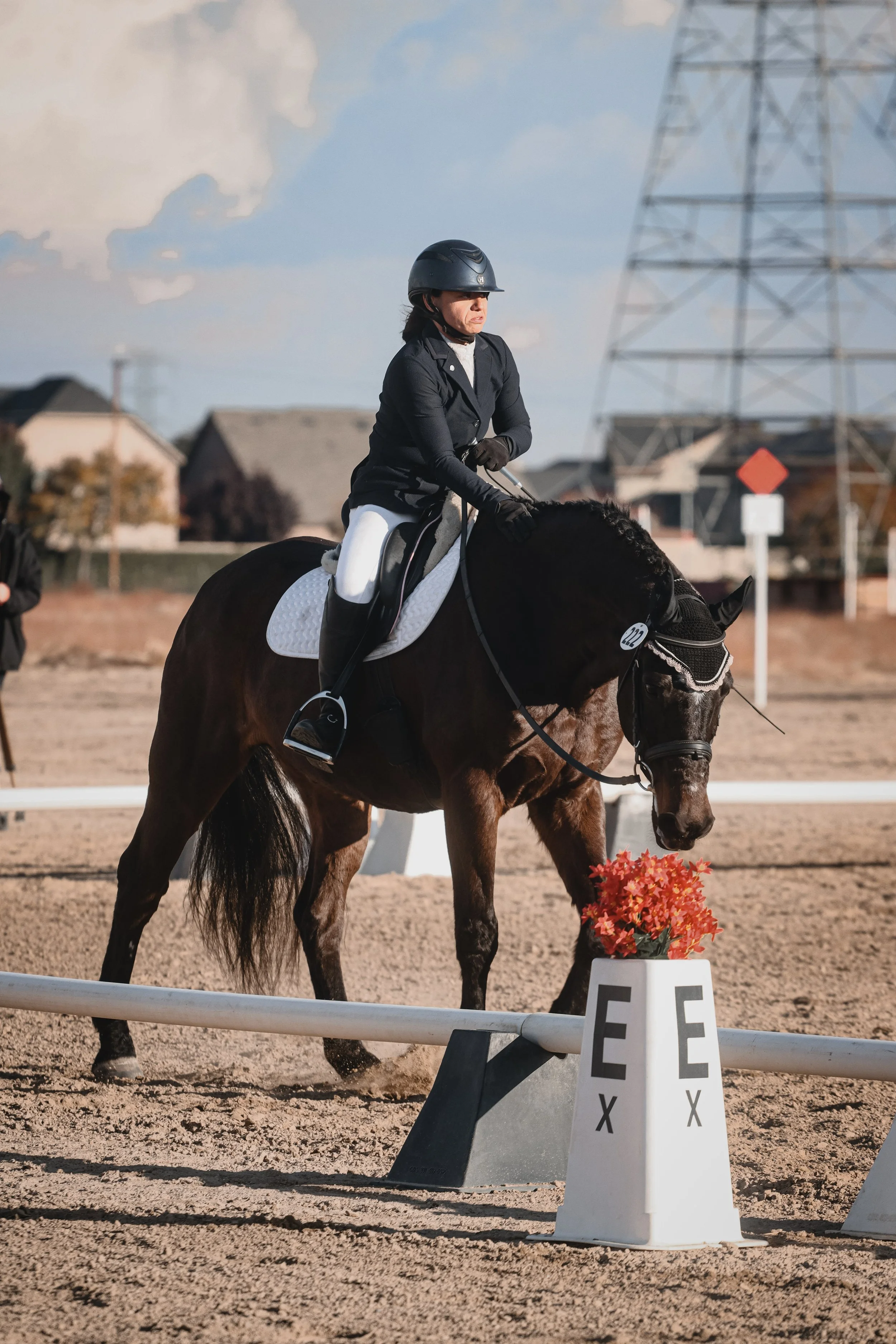 A woman riding a brown horse in an equestrian arena, wearing a helmet, black jacket, white riding pants, and gloves. There is a flower arrangement and a sign with the letter F in the foreground, and a large metal power line tower in the background.