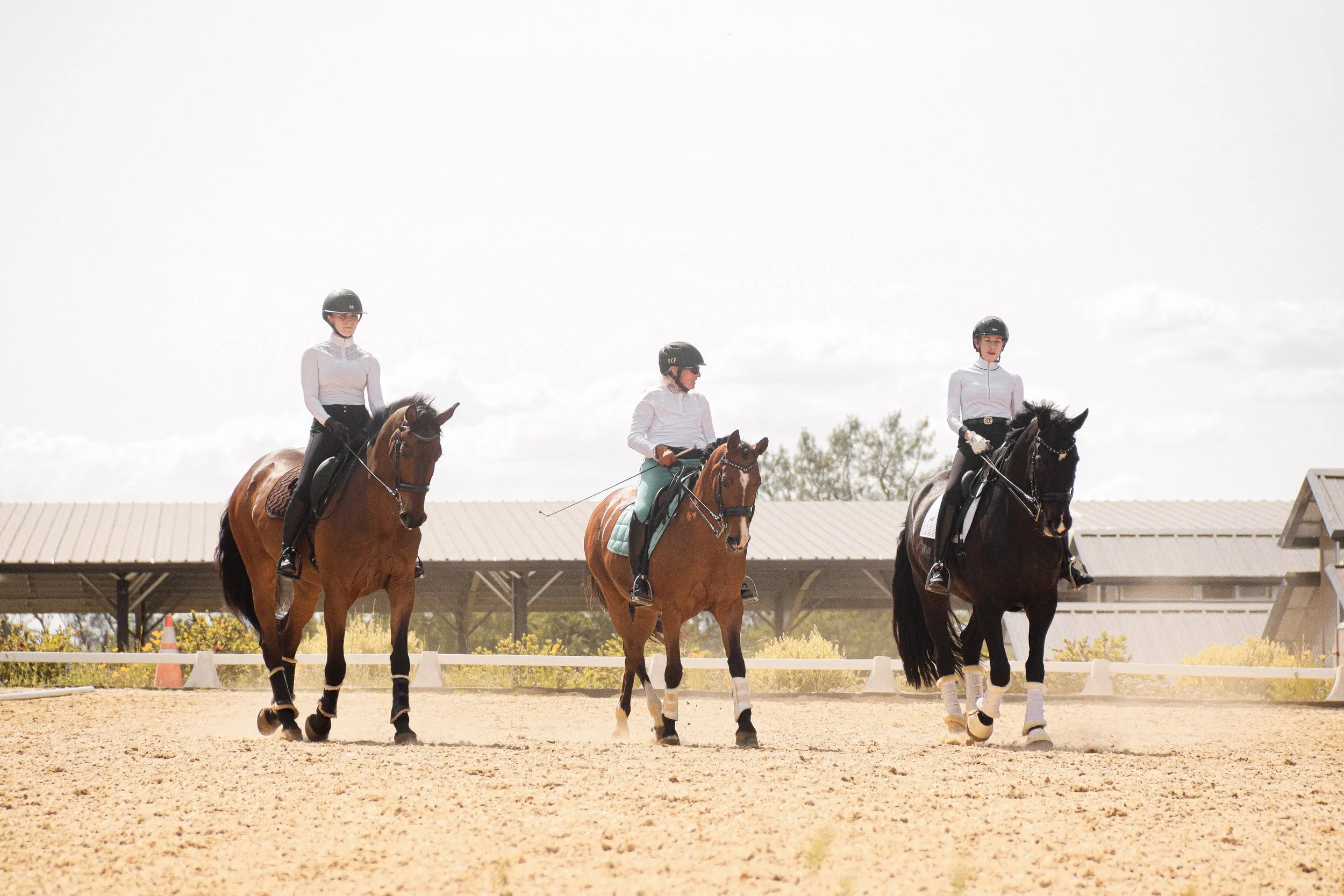 Three female horseback riders in white riding attire and helmets riding on a sandy outdoor arena with a beige roof structure in the background.