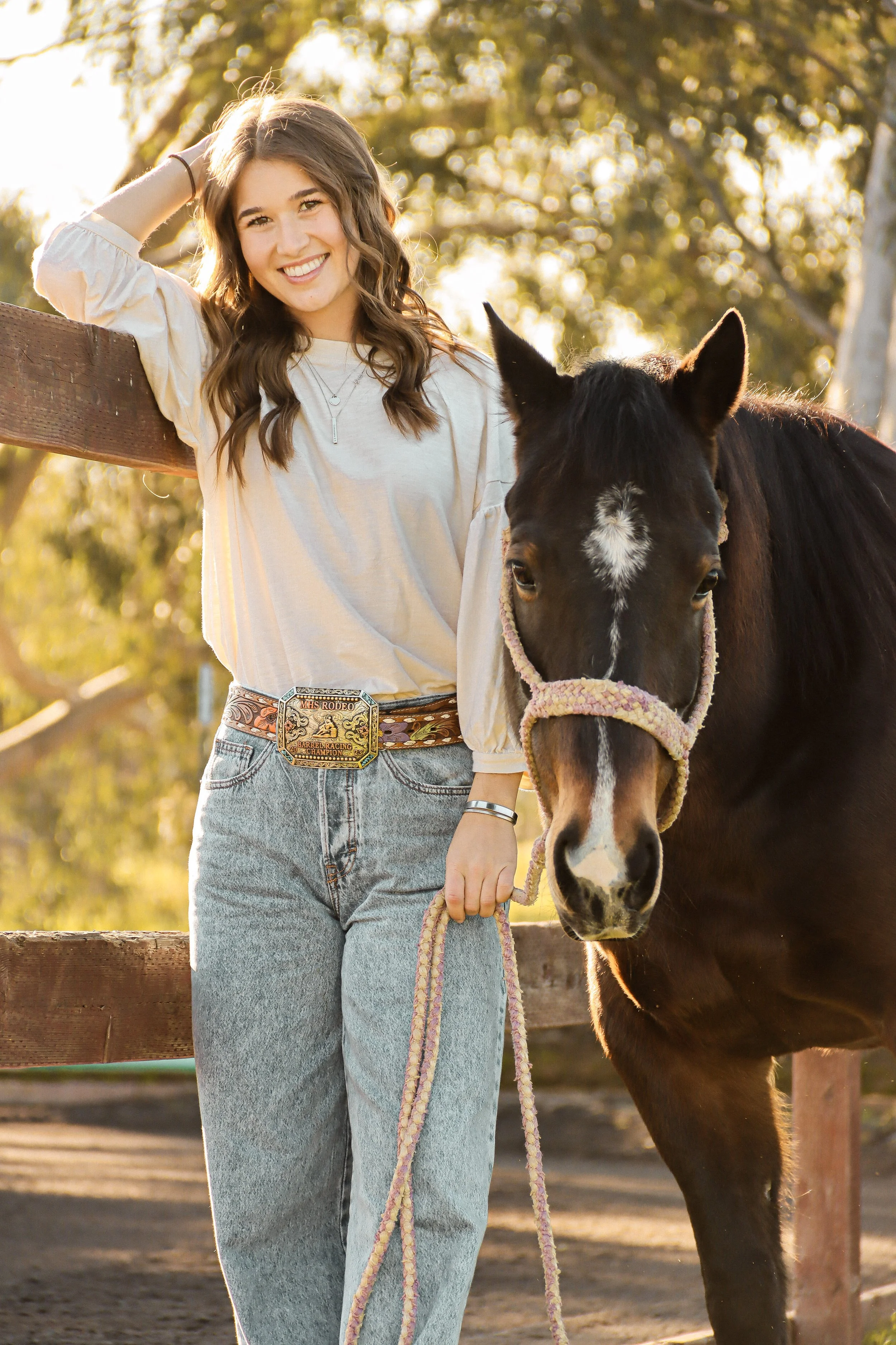 A young woman with long brown hair smiling and standing outdoors with a black horse, holding a lead rope, during sunset on a farm or rural area.