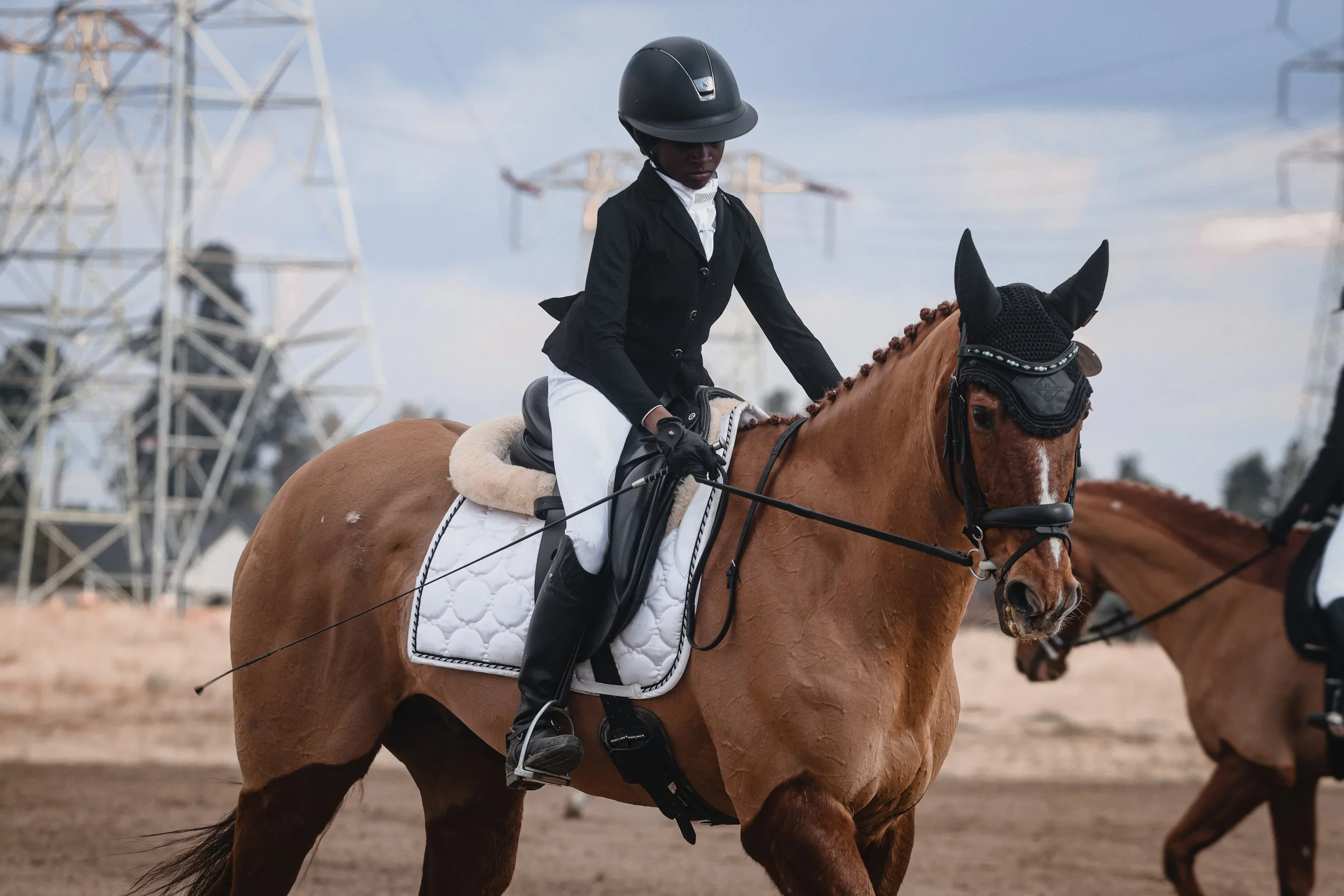 A rider dressed in a black riding jacket, white breeches, and a black helmet riding a brown horse with a braided mane, in an outdoor equestrian arena with power lines and a blue sky in the background.