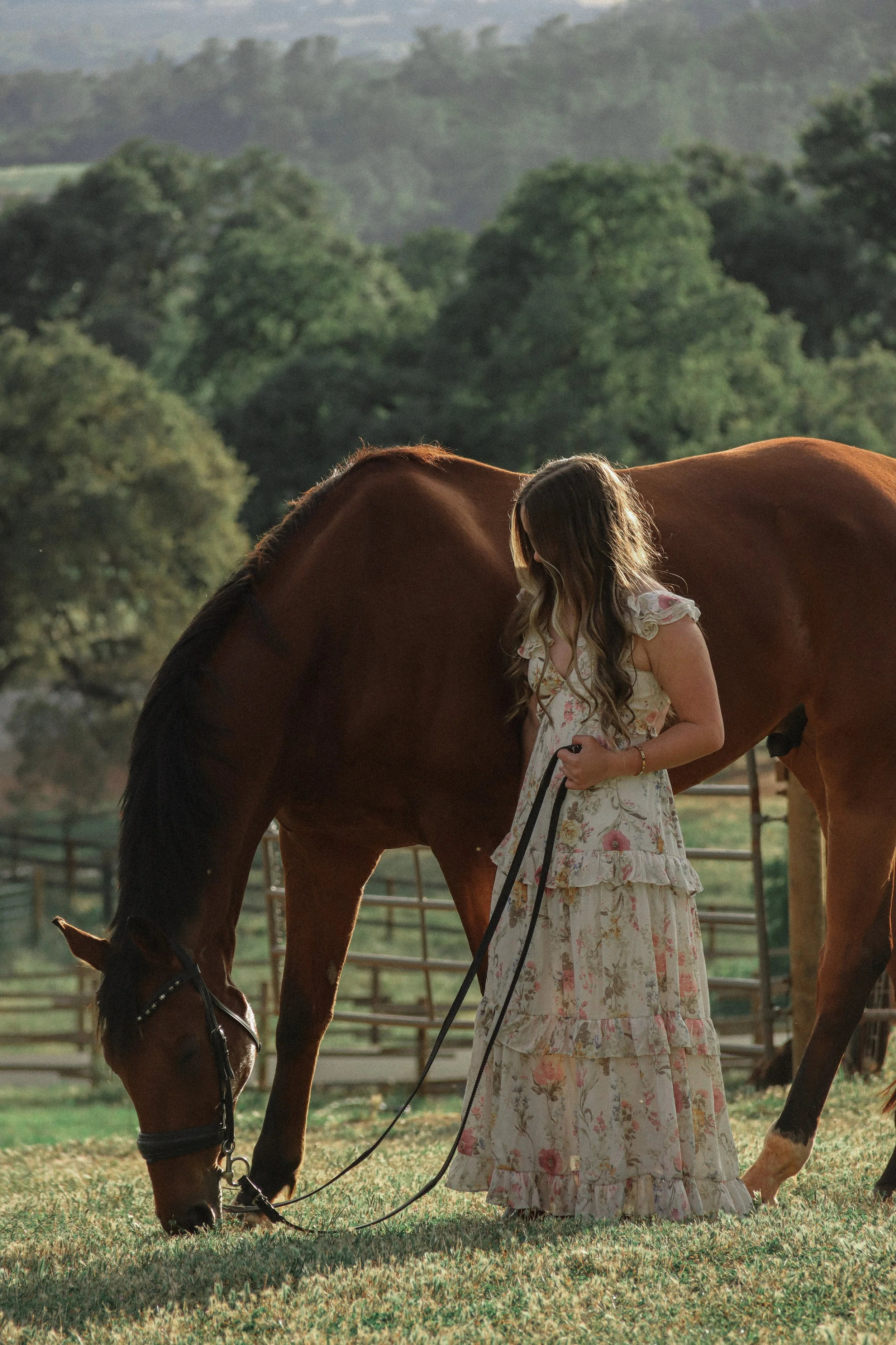 A woman with long wavy hair in a floral dress standing next to a brown horse, holding its reins, in a green outdoor setting with trees and hills in the background.