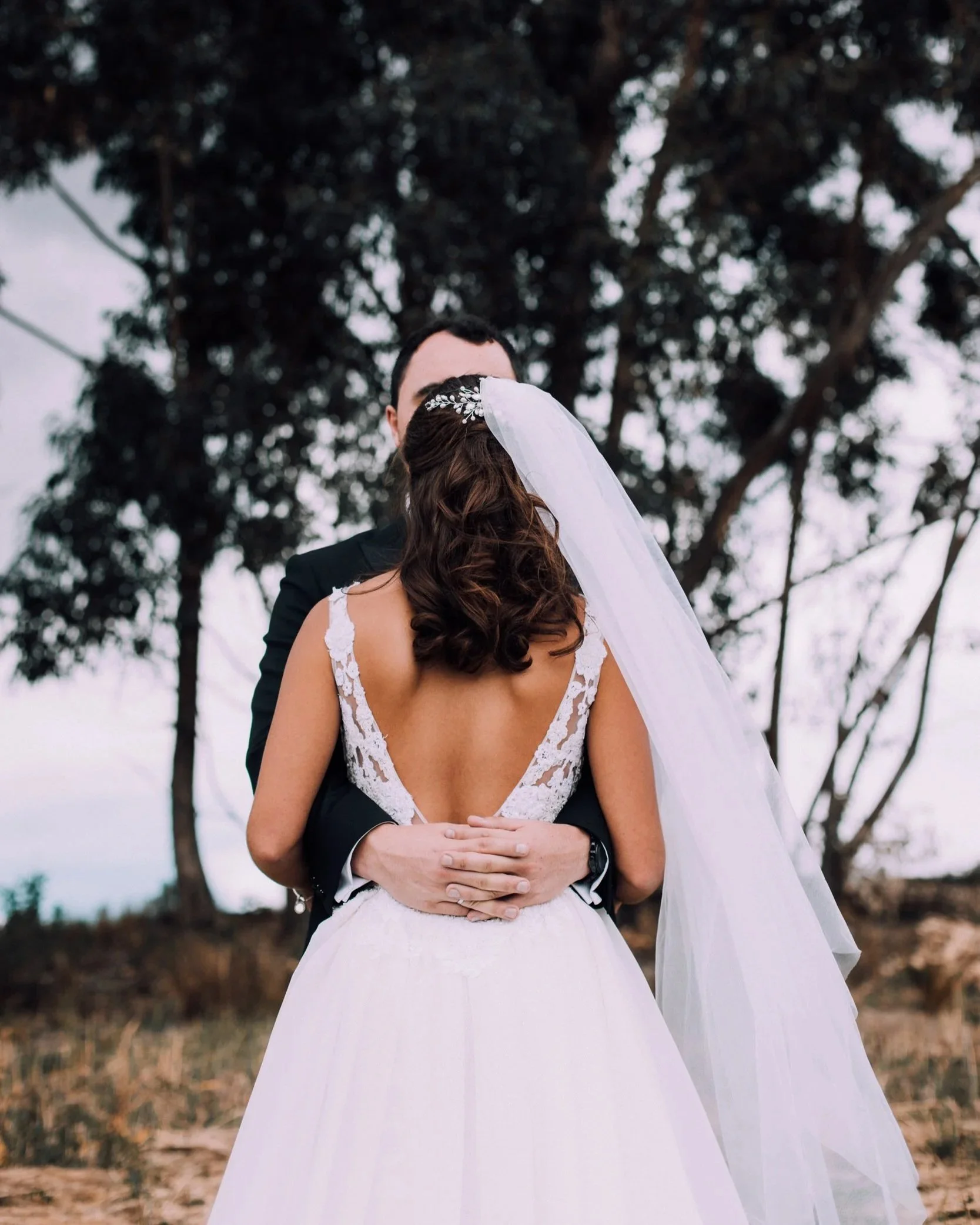 A couple in wedding attire sharing a close embrace outdoors, with a tree and cloudy sky in the background. The bride's back and veil are visible, with the groom's face partially obscured.