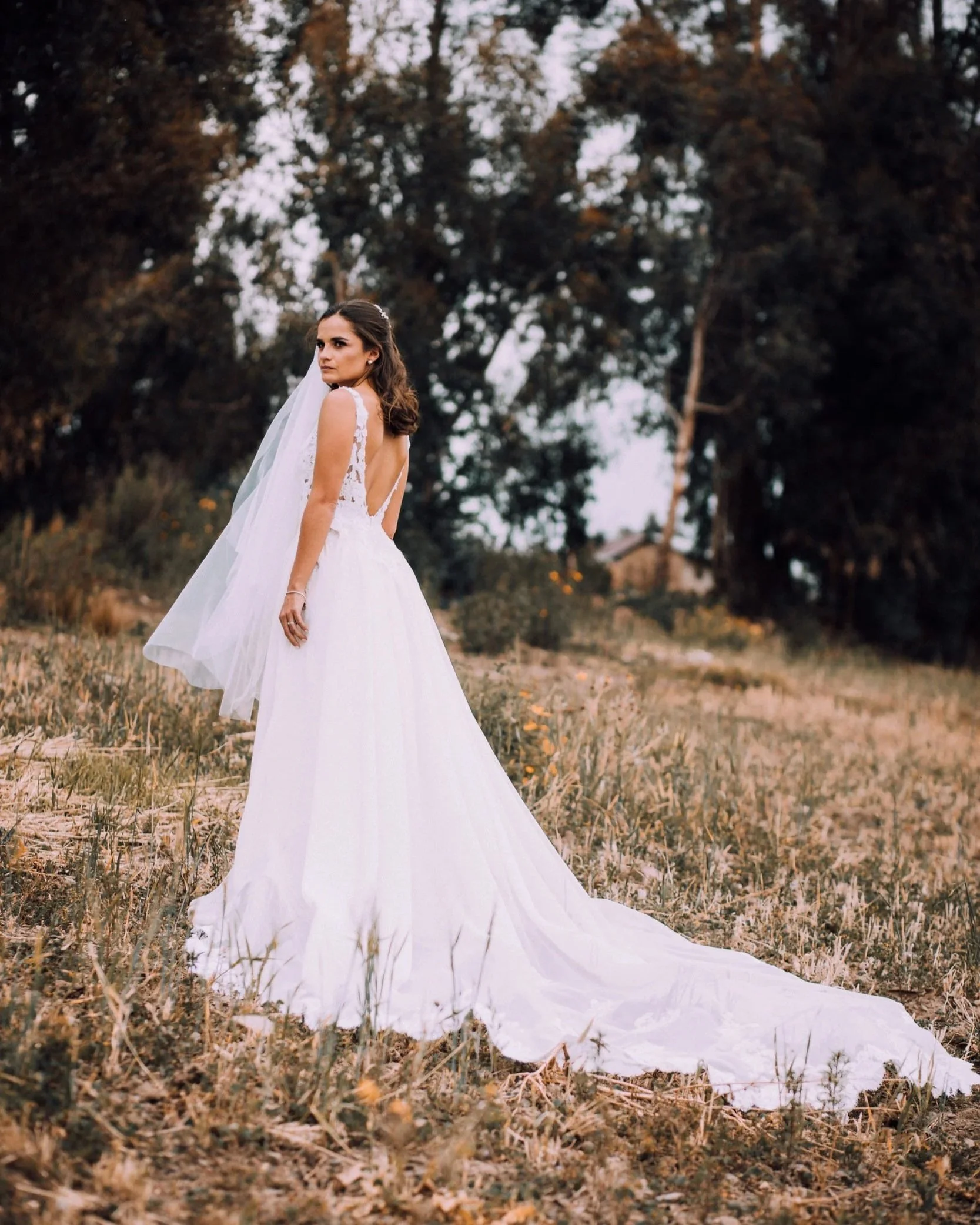 A woman in a wedding dress standing outdoors on a grassy field with trees in the background.