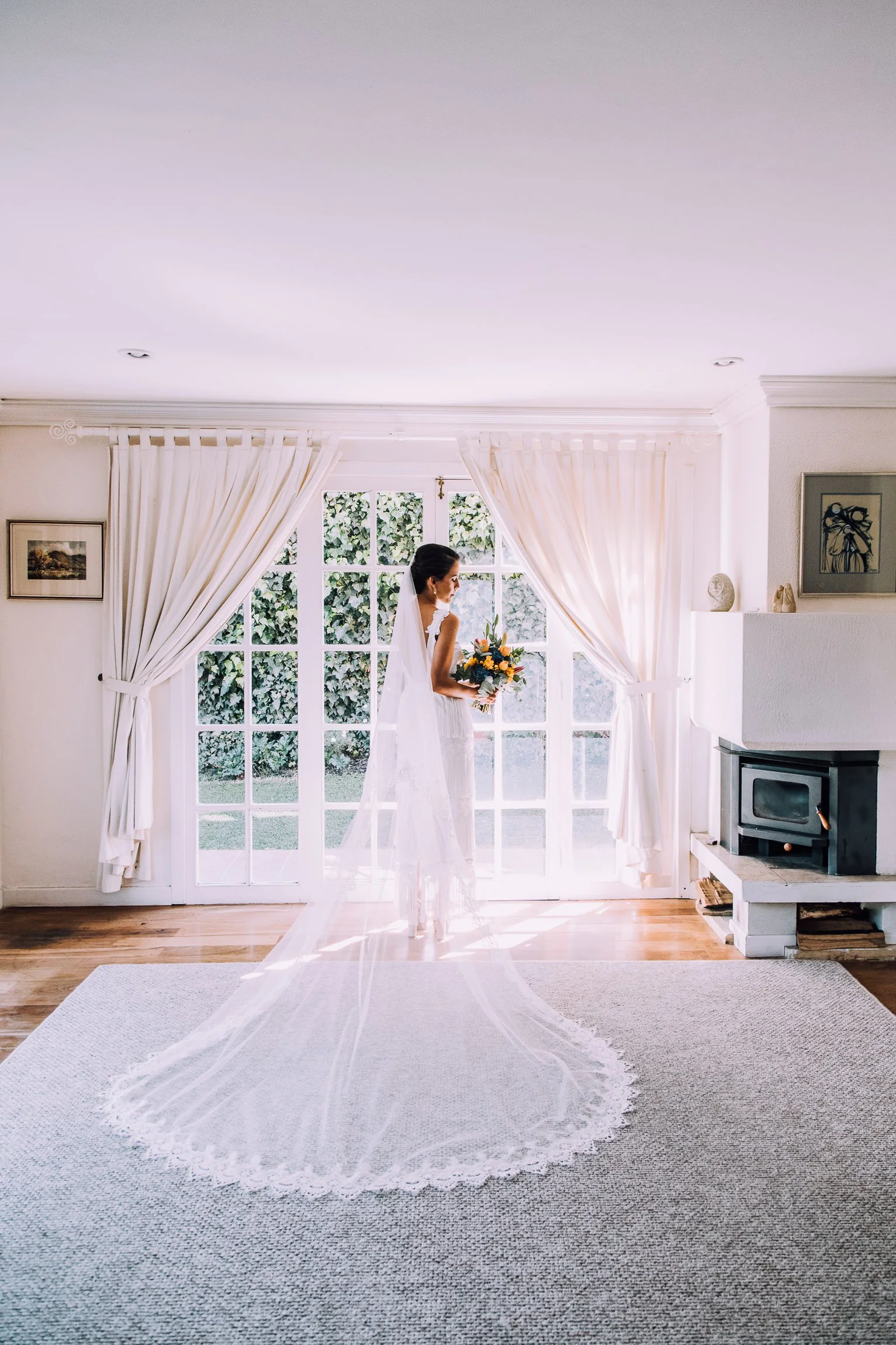 A bride in a wedding dress holding a bouquet standing in front of a large window with curtains, with a wood stove and framed artwork in the background.