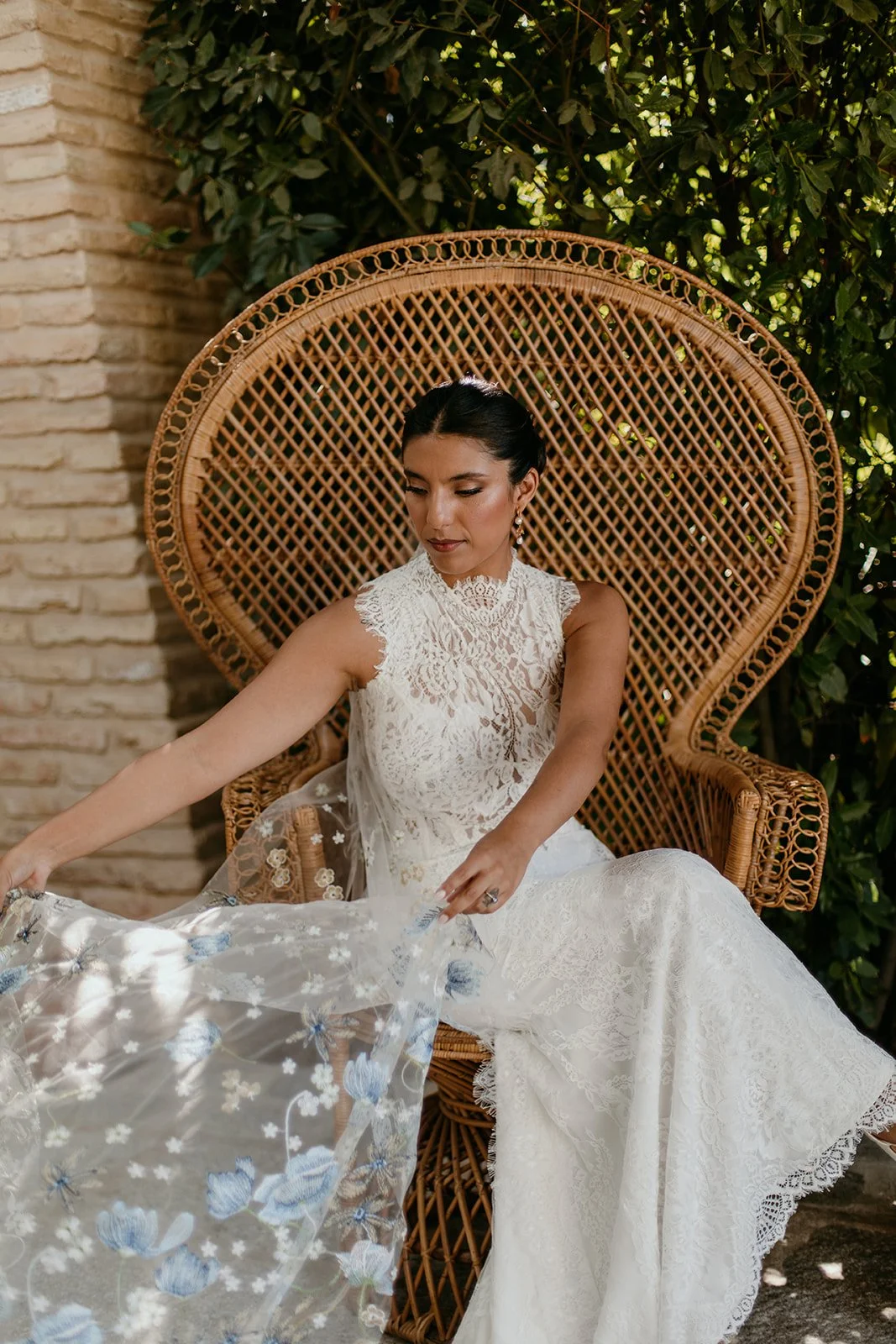 A woman dressed in a white lace wedding gown sitting on a rattan peacock chair, fans her veil which has blue flora embroidered throughout. Outdoors with green foliage and a brick wall in the background.