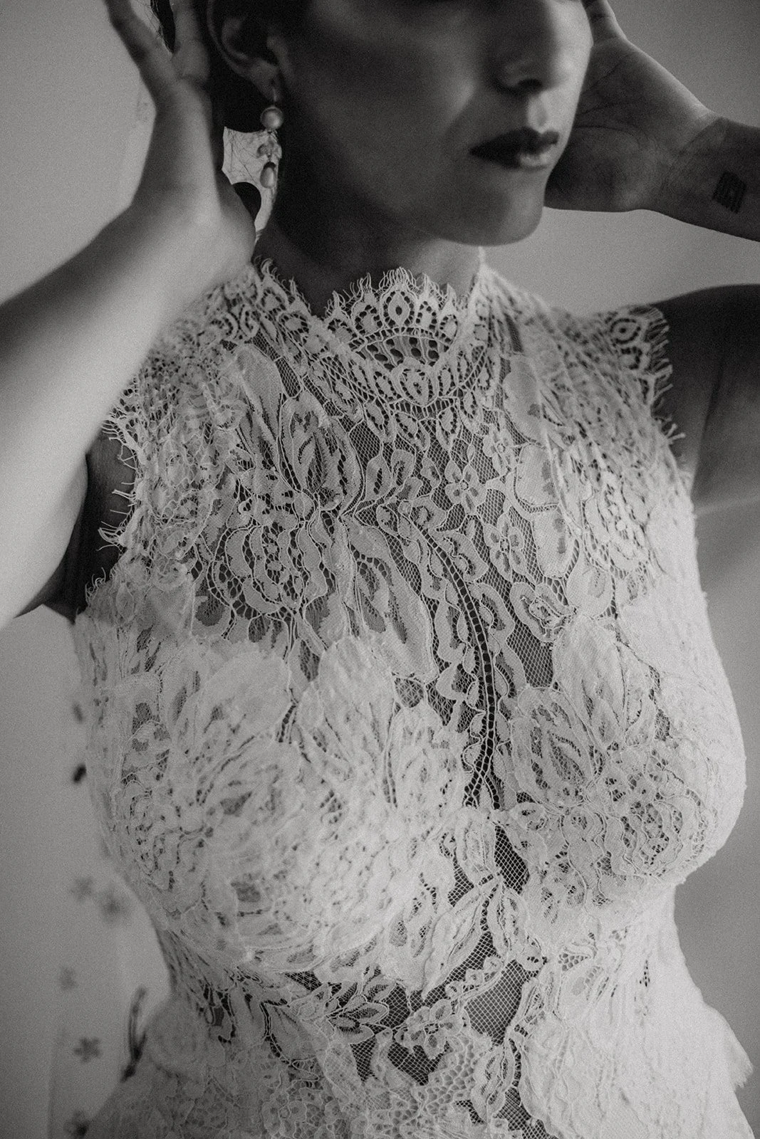 Close-up of a woman wearing a sheer lace dress with floral patterns, earrings, and dark hair, with her hands near her head.