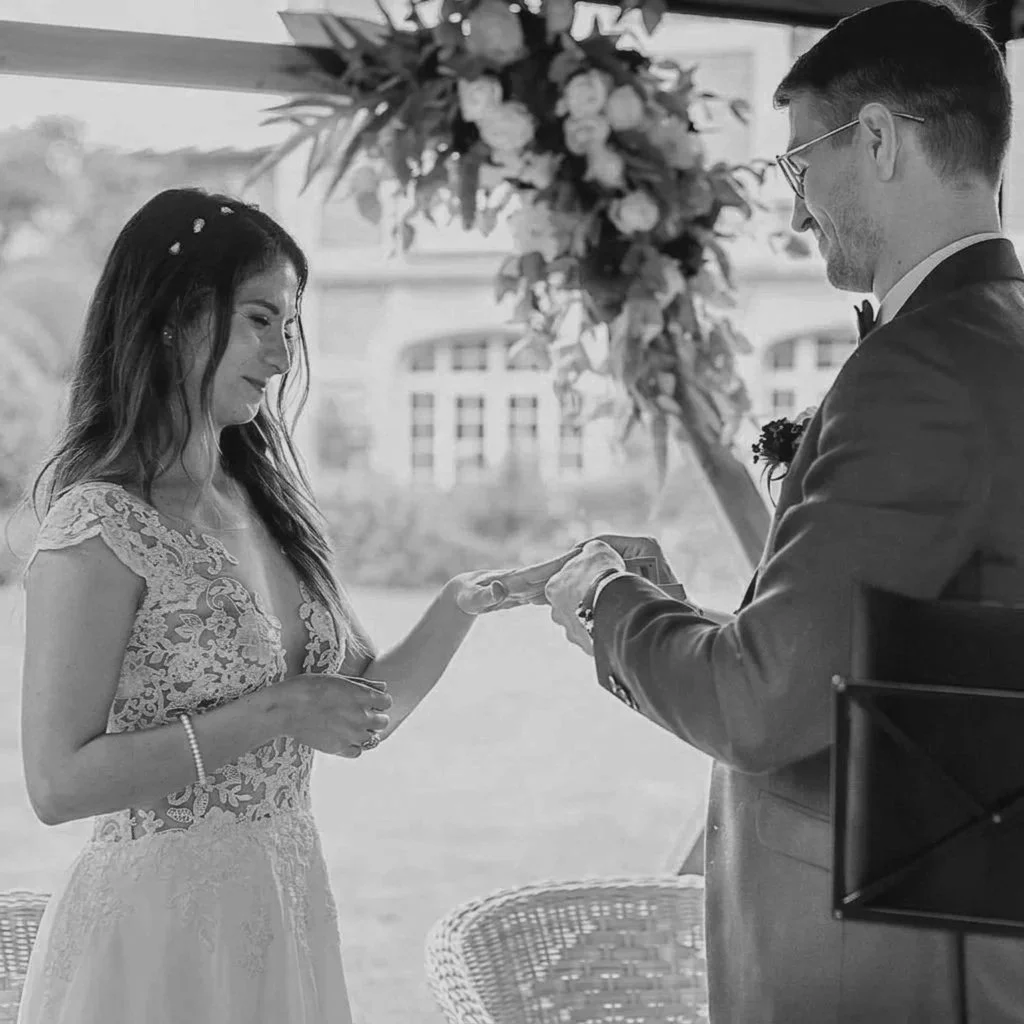 A bride and groom exchange rings during their wedding ceremony, with a floral arrangement and outdoor backdrop visible.