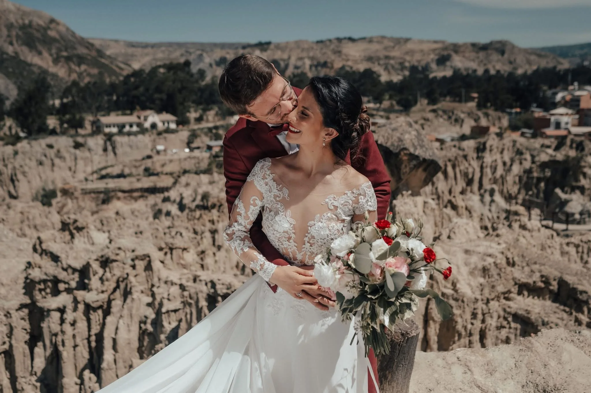 A bride and groom sharing a kiss outdoors with a mountainous landscape in the background. The bride is holding a bouquet of white and red flowers, and she is wearing a lace wedding dress. The groom is dressed in a red suit.