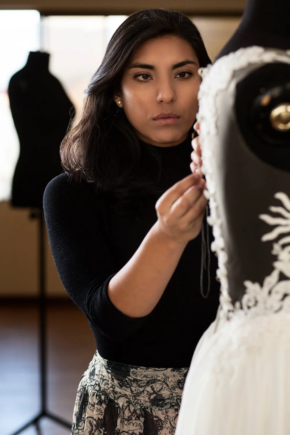 A young woman with dark hair wearing a black top and patterned skirt, adjusting a wedding dress on a mannequin in a fashion design studio.