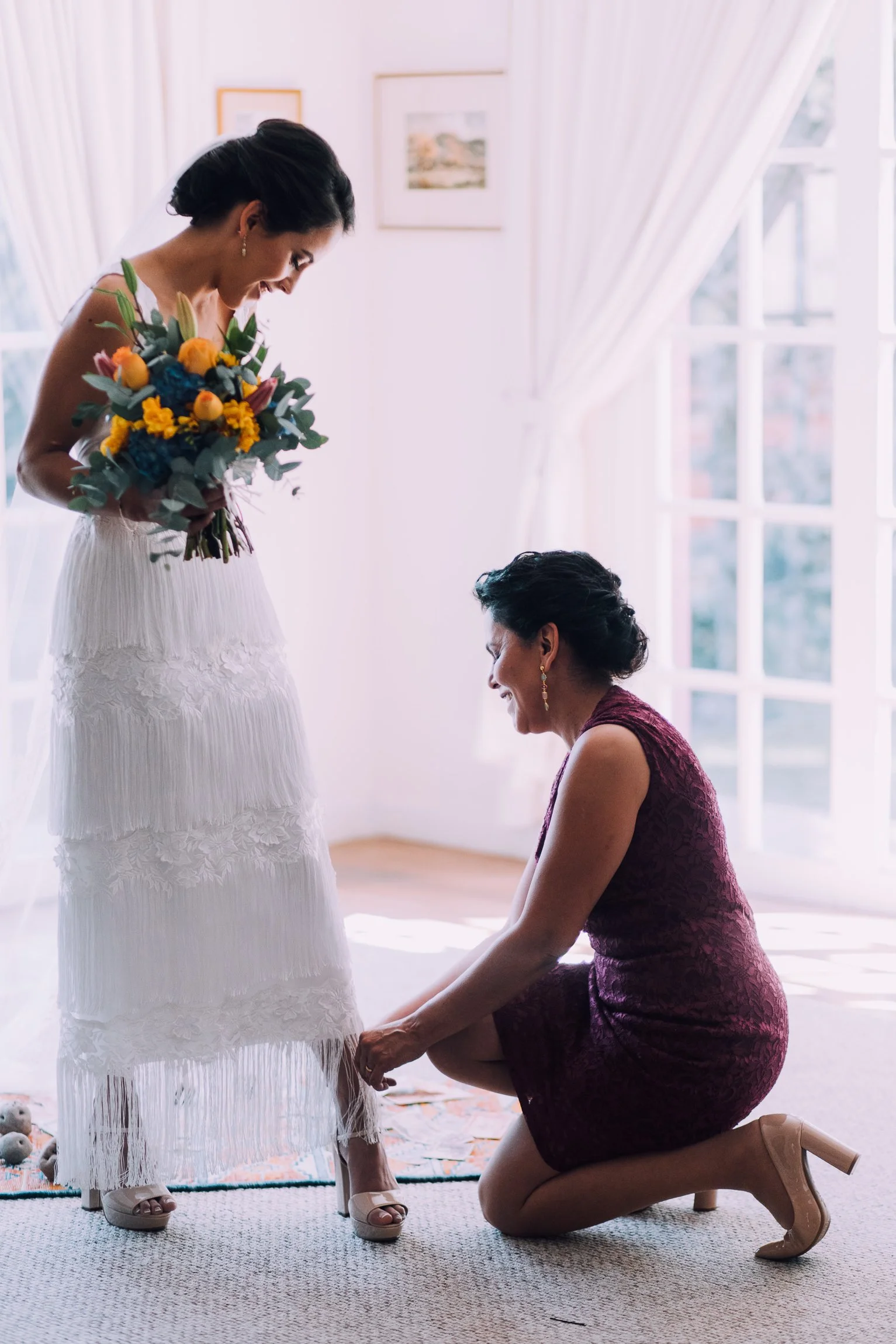 A bride in a white wedding dress holding a bouquet of flowers stands as an older woman helps her buckle her heels, holding the bride's hands, in a sunlit room with large windows and white curtains.