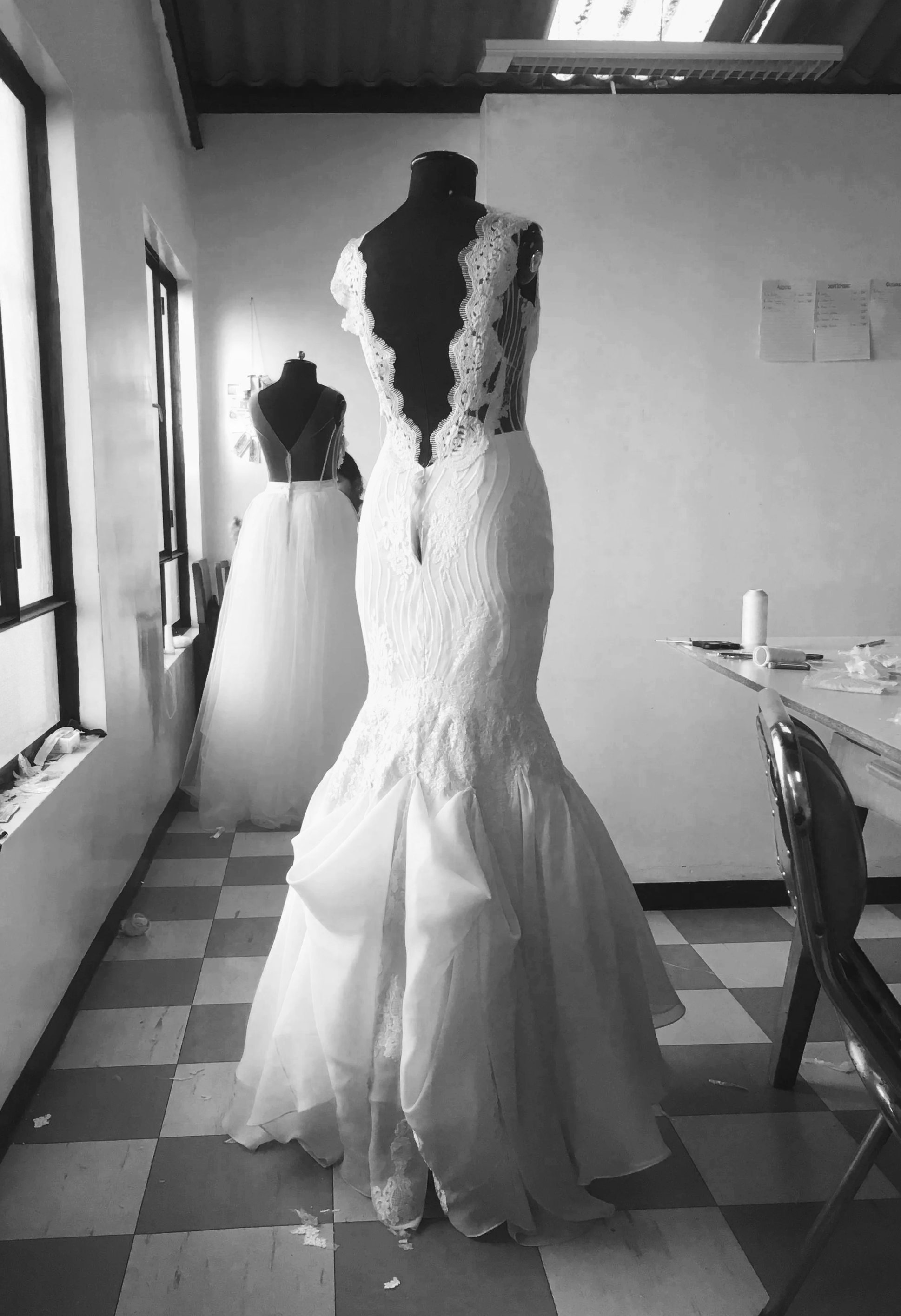 A wedding dress on a mannequin in a room. The dress is fitted, with lace details, and a long train. There is another mannequin in the background with a similar dress. The room has a window, some chairs, and sewing materials on the table.