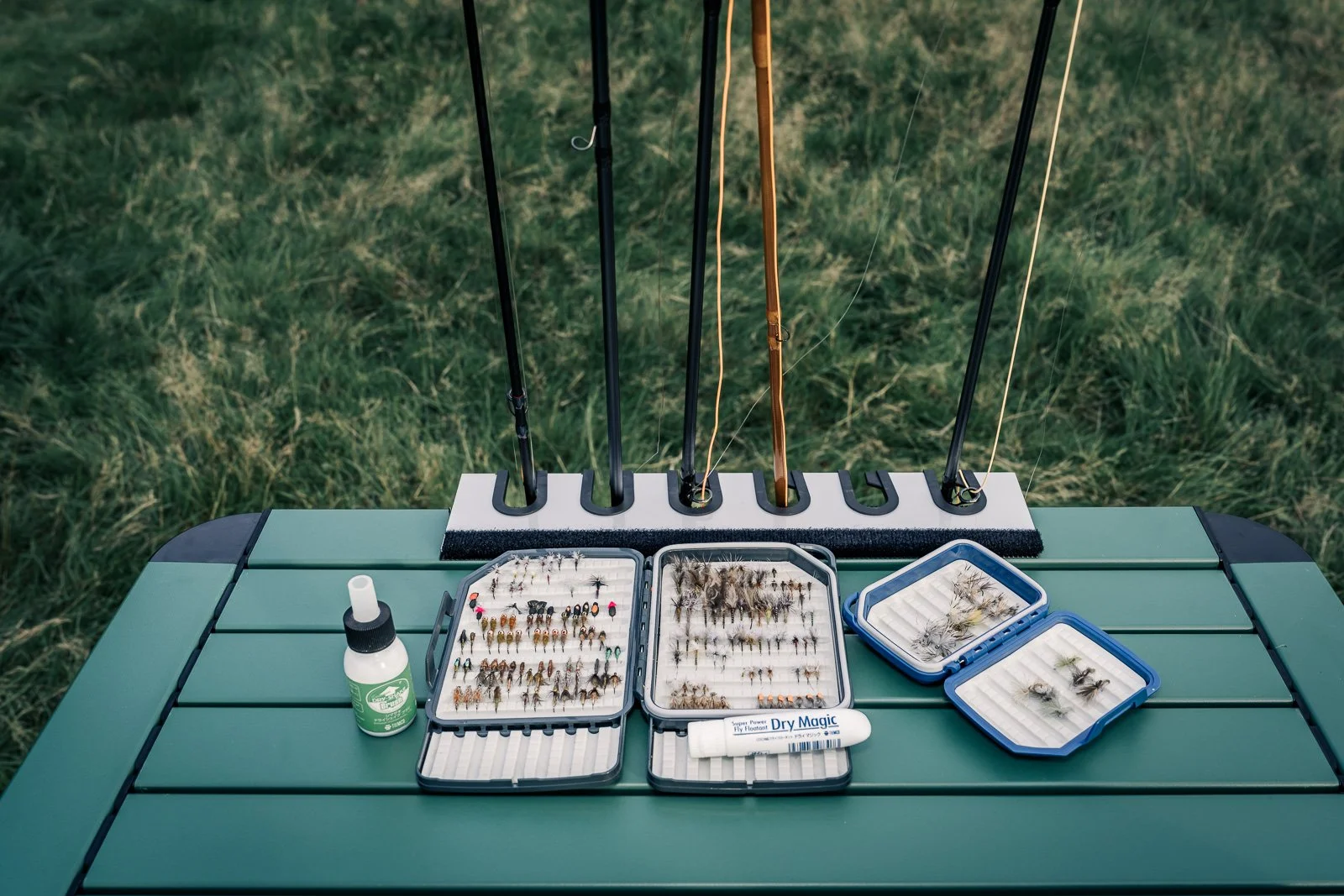 Fly-fishing rods, assorted fly boxes, fishing flies, and fly floatant arranged on a green outdoor table.