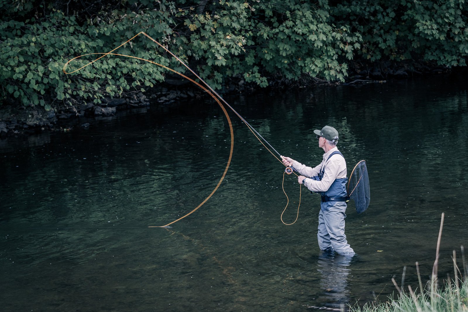 Fly fisherman wading in a river and casting a fly line near a forested bank