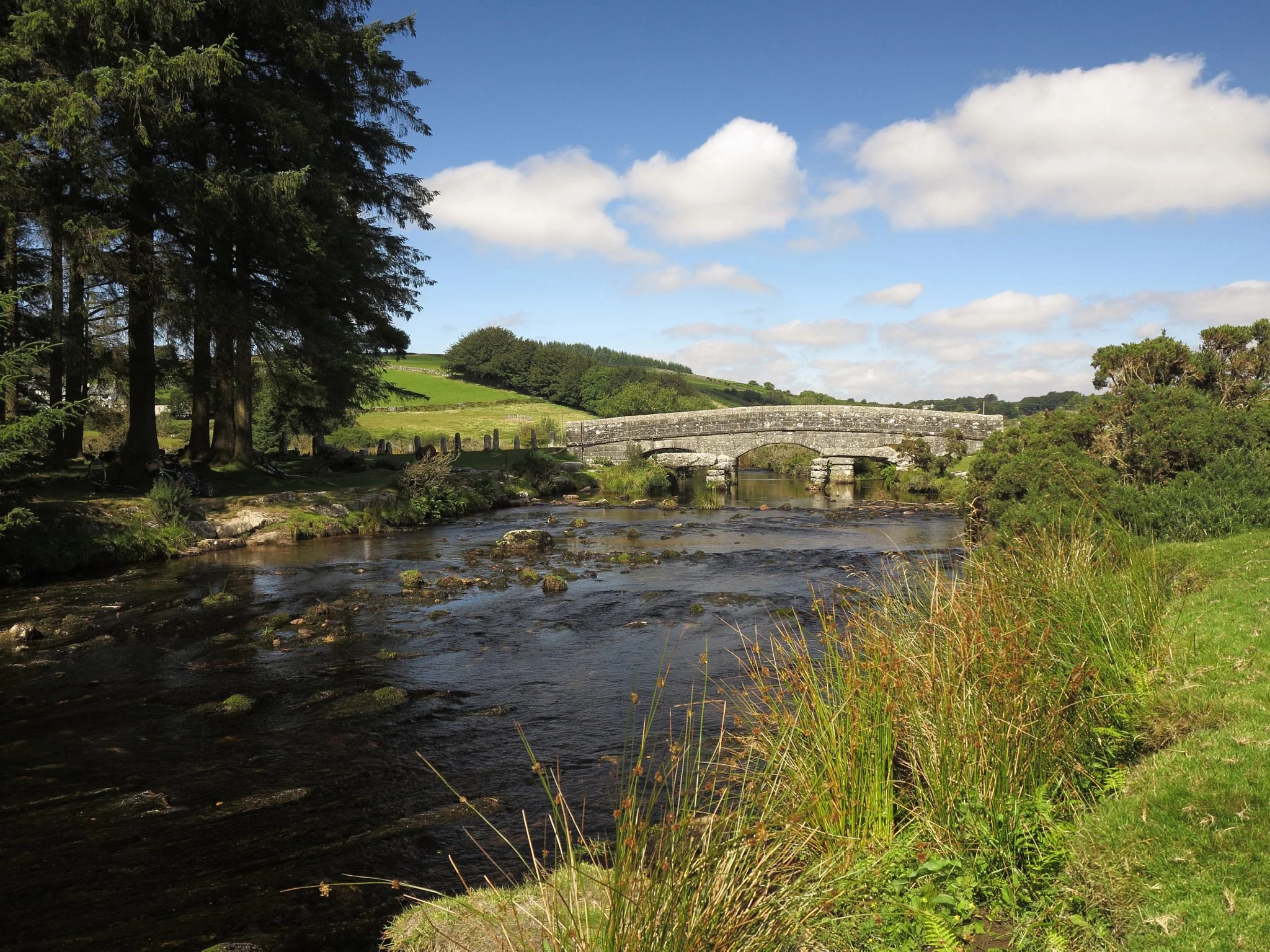 Historic stone arch bridge crossing a shallow river in a green countryside landscape under a blue, cloud-filled sky