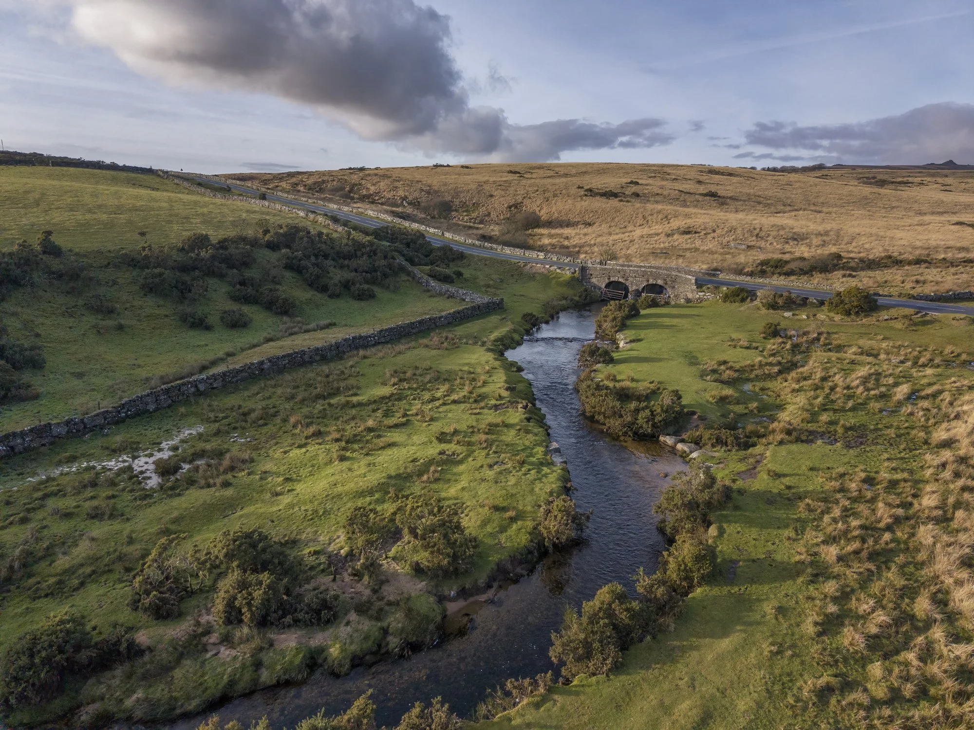 Aerial view of a stone-arched bridge carrying a rural road over a winding river through grassy moorland beneath cloudy skies