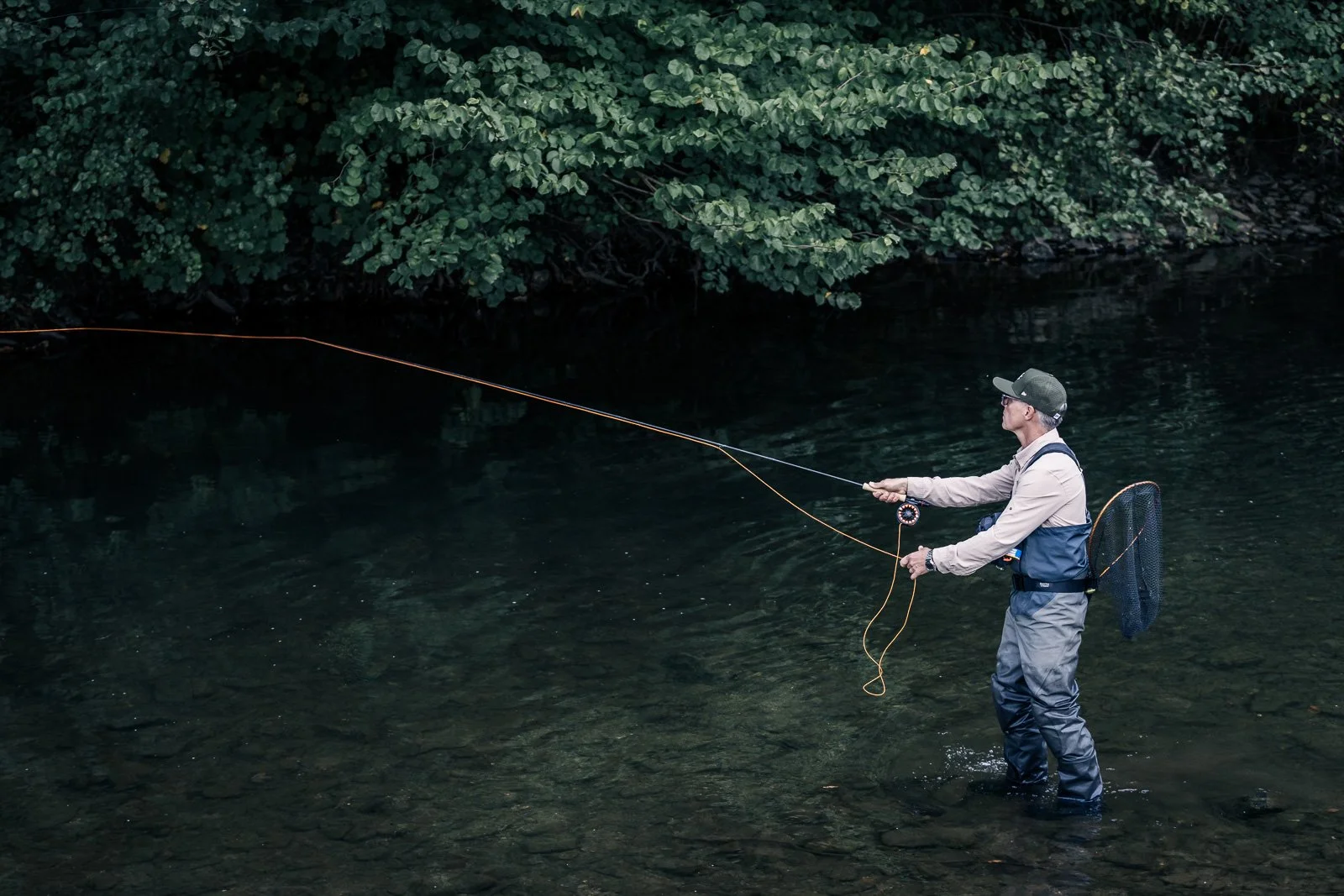 Fly fisherman wading in a clear river beneath leafy trees