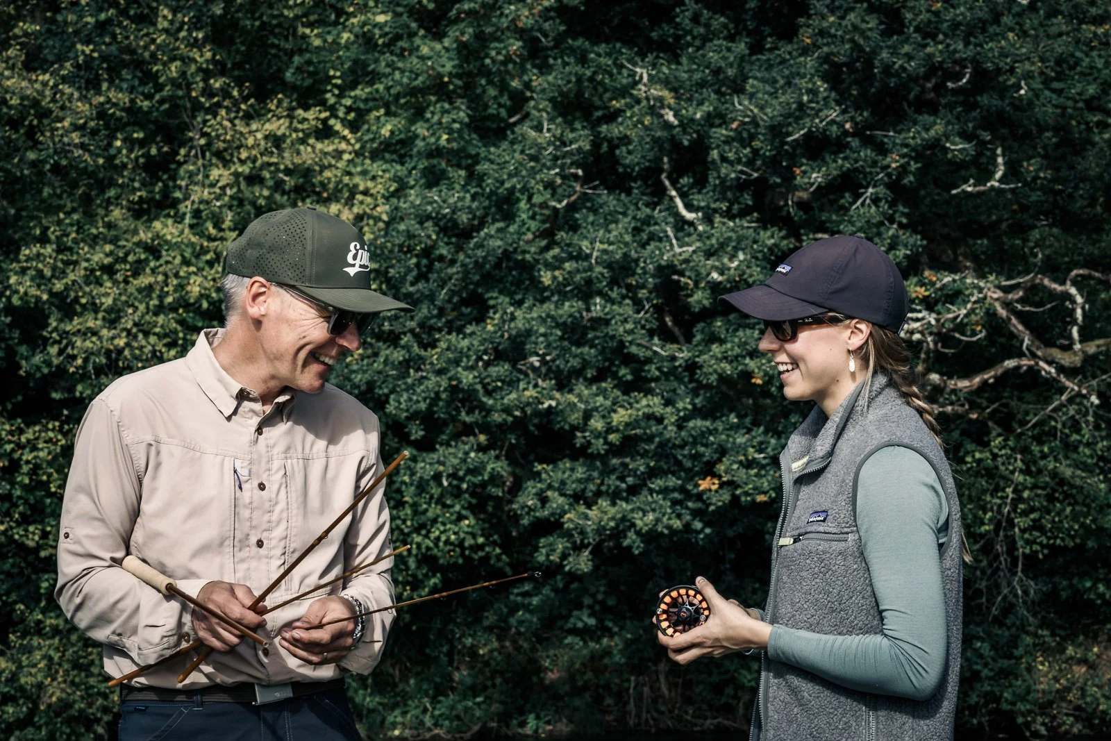 Two fly-fishing enthusiasts wearing sunglasses and caps talk outdoors in a wooded setting, holding fishing rods and a fly reel.