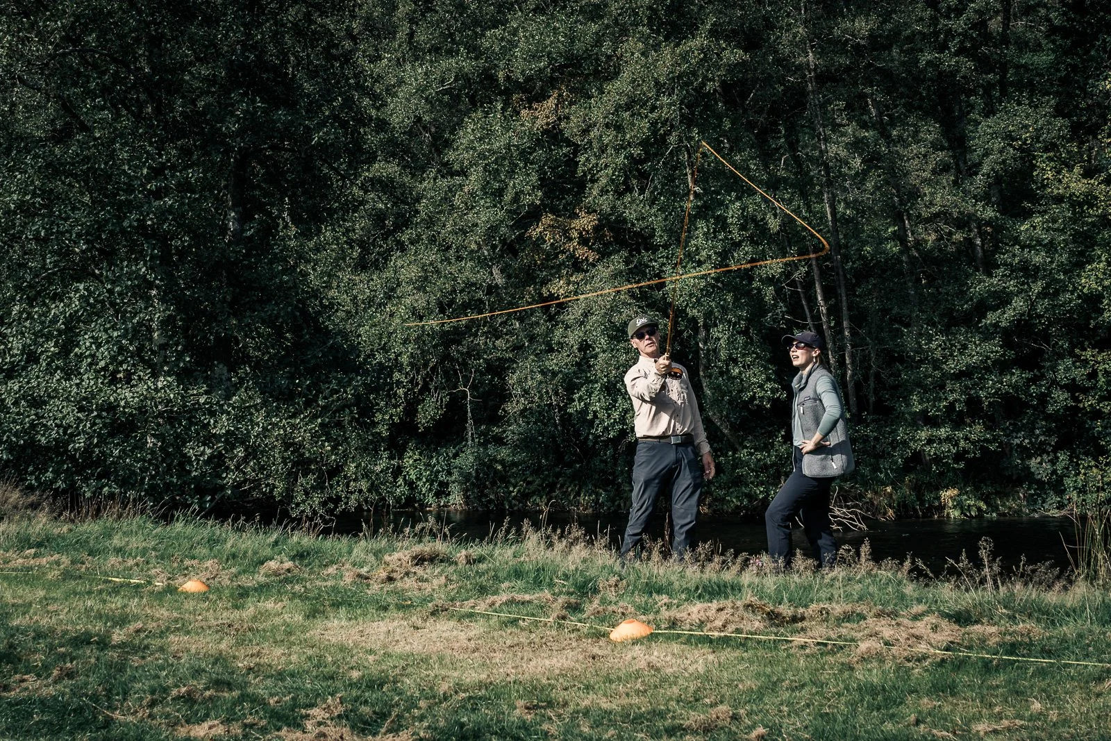 Two people fly fishing beside a wooded stream, with a fishing line cast across the clearing.