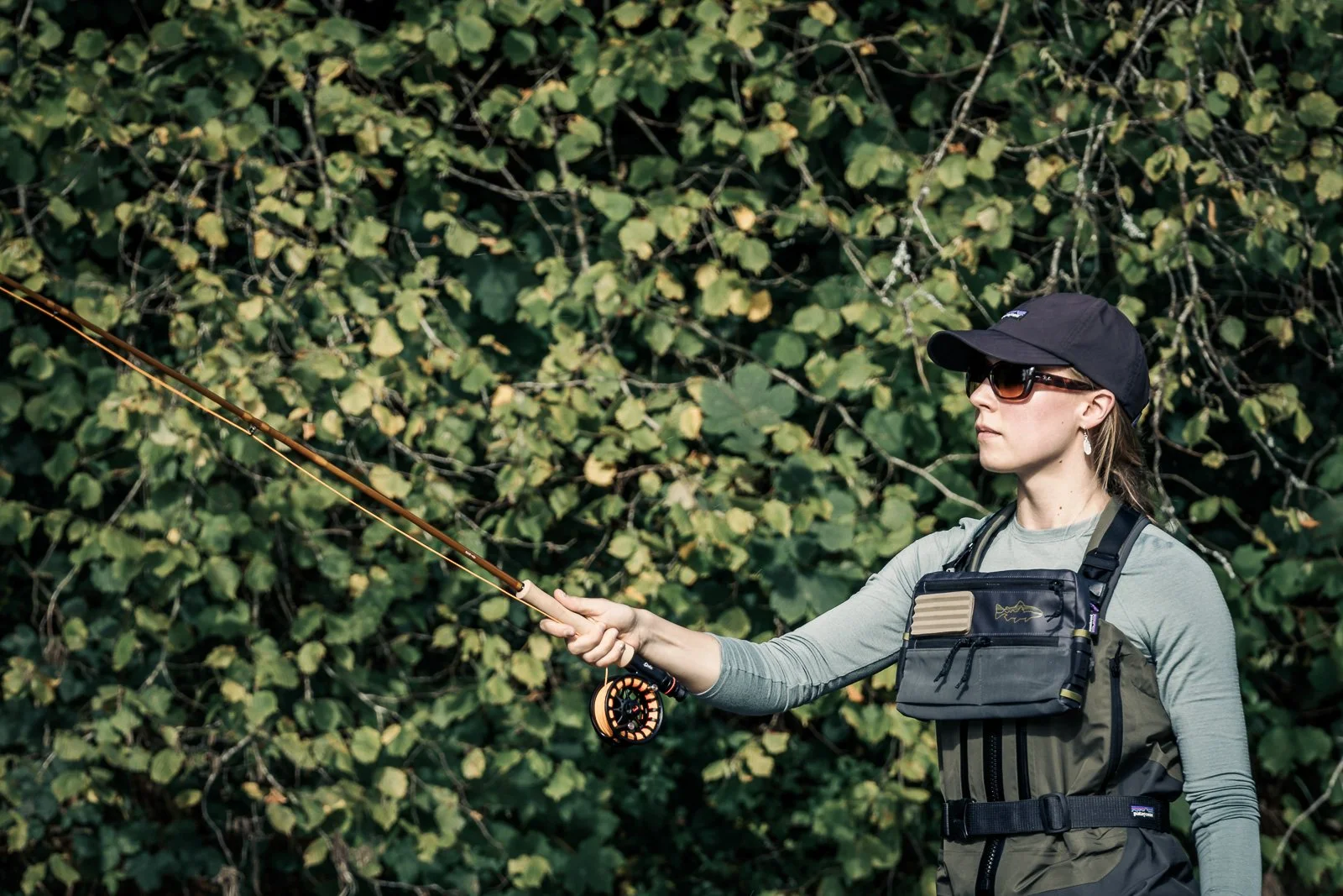 Fly fisher wearing sunglasses and waders casts a fishing rod in a wooded area.