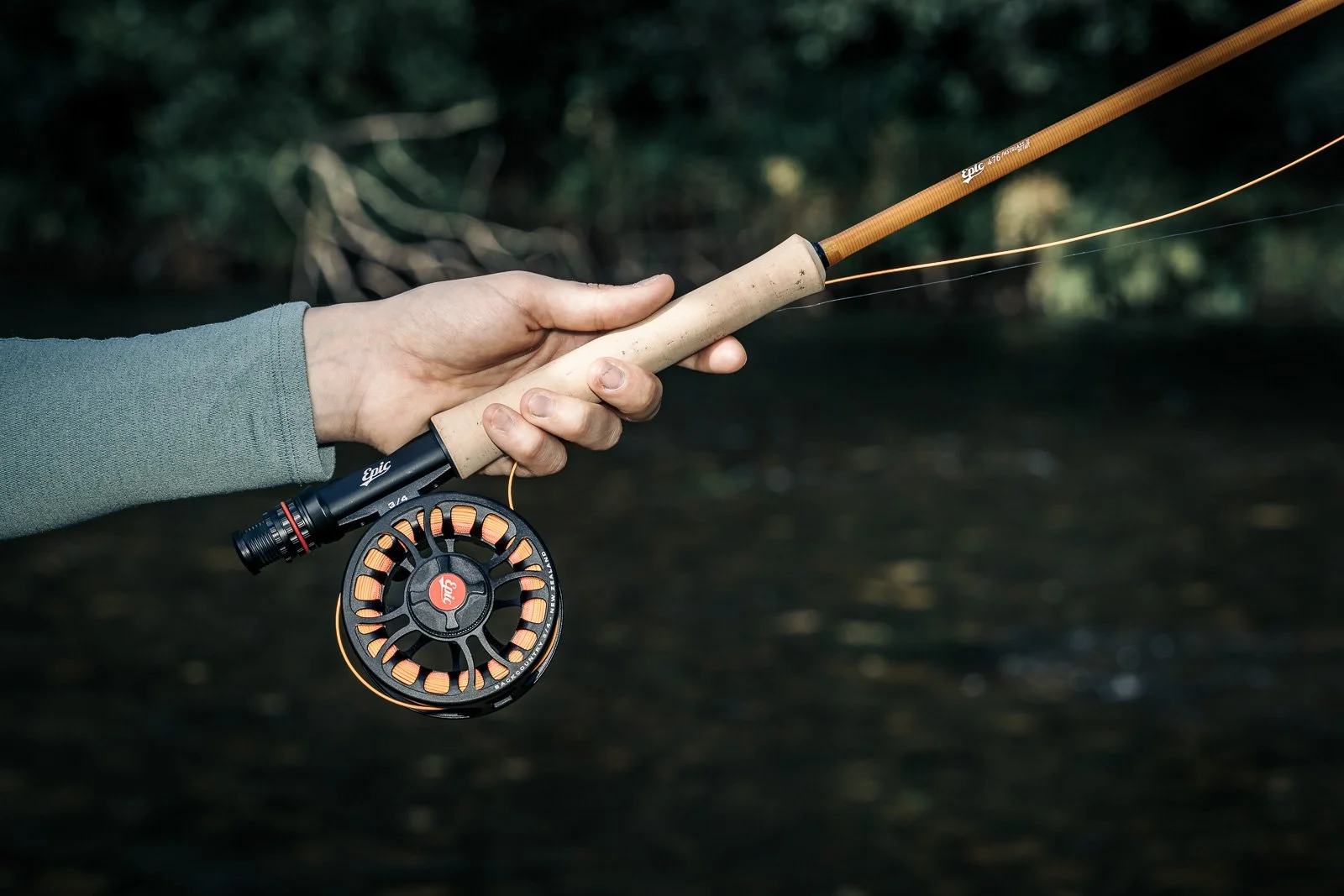 Close-up of a hand holding an Epic fly-fishing rod and reel over a shaded stream