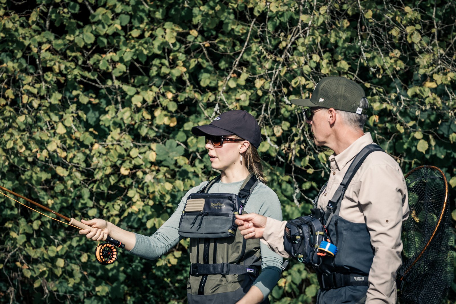 Two fly fishers in waders holding a fly rod and net outdoors in a wooded area