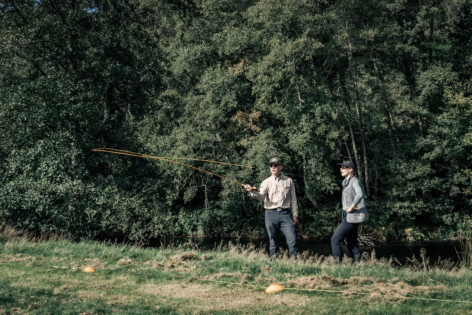 Two people fly-fishing beside a wooded stream, with orange fly lines extending over the water.