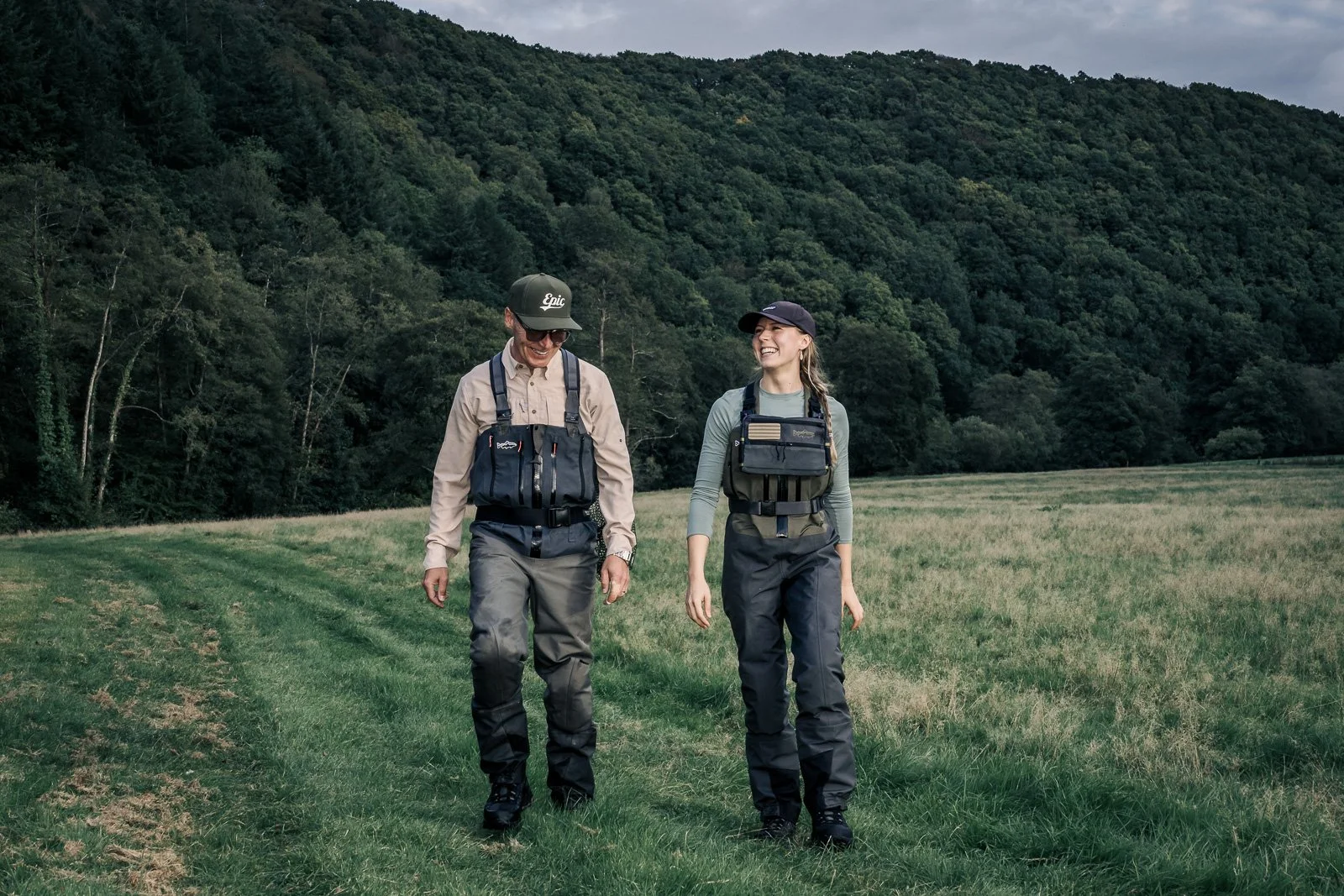 Two smiling anglers in chest waders and fishing vests walking through a grassy meadow with a forested hillside behind them