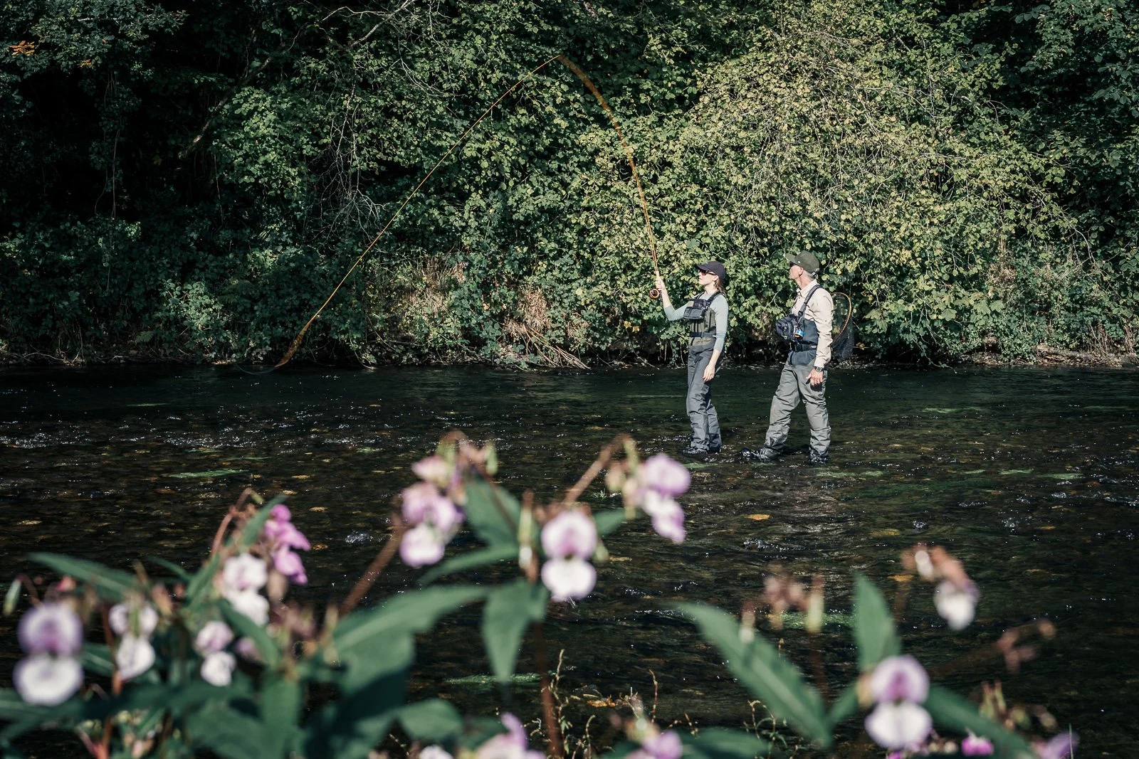 Two anglers fly-fishing in a forest stream, framed by wildflowers.