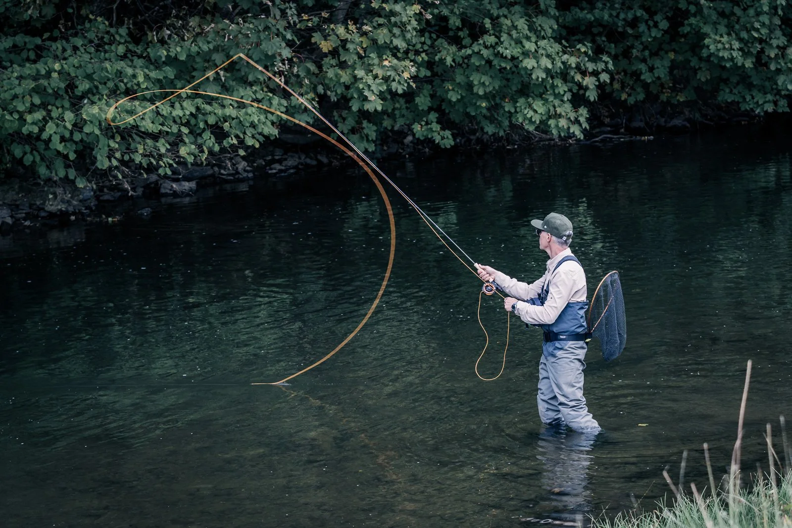 Fly fisherman wading in a river and casting a line beneath a forested riverbank