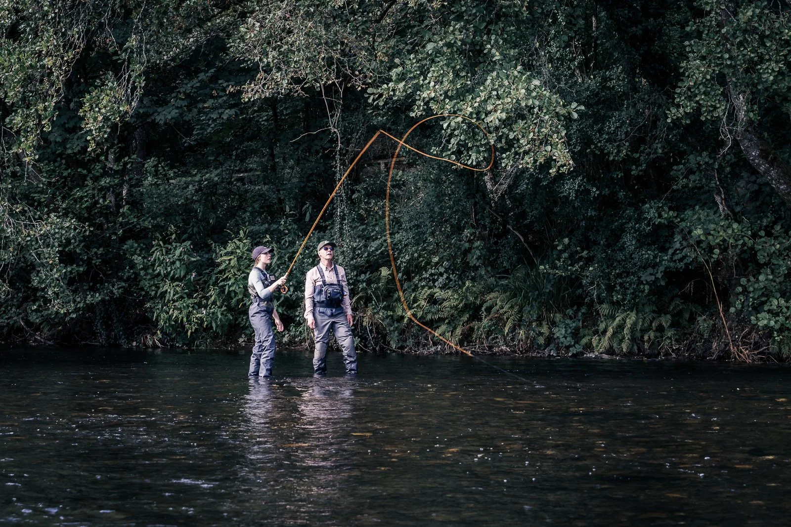 Two fly fishers wading in a forest river, casting an orange fly line.