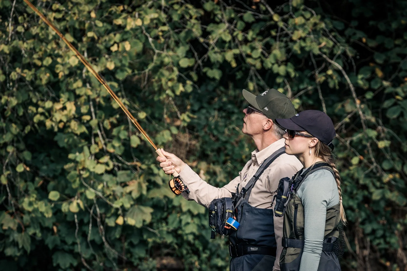 Two fly-fishers wearing waders and sunglasses cast a fishing rod in a wooded area
