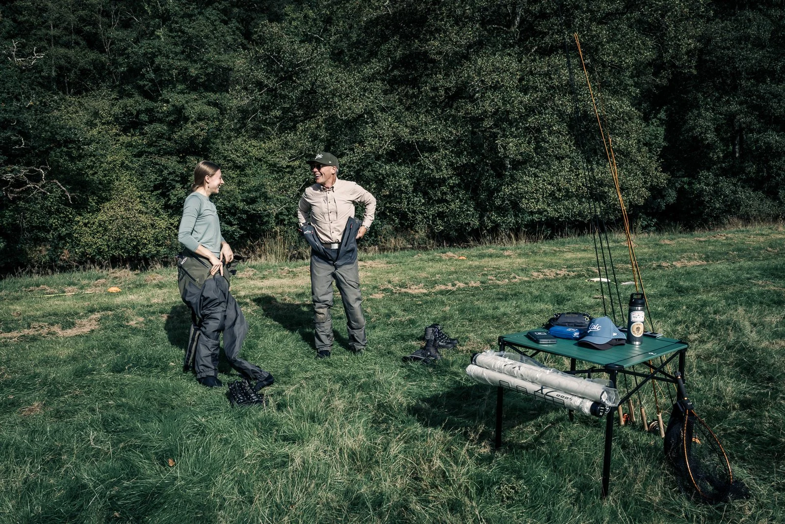 Two fly fishers prepare outdoors beside a table holding fly-fishing rods, gear, and a landing net.