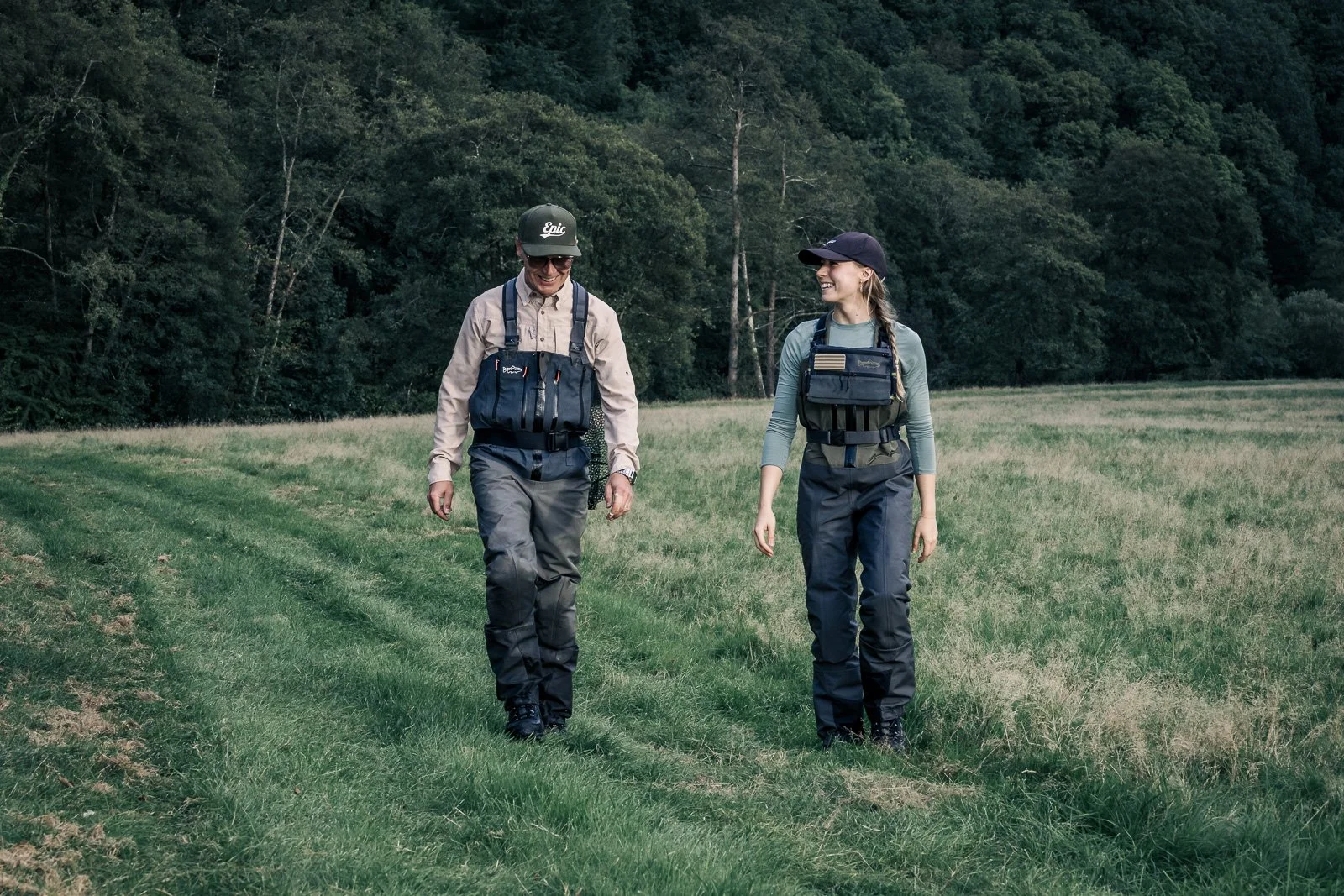 Two anglers wearing chest waders and fishing vests walk through a grassy field beside a forest.