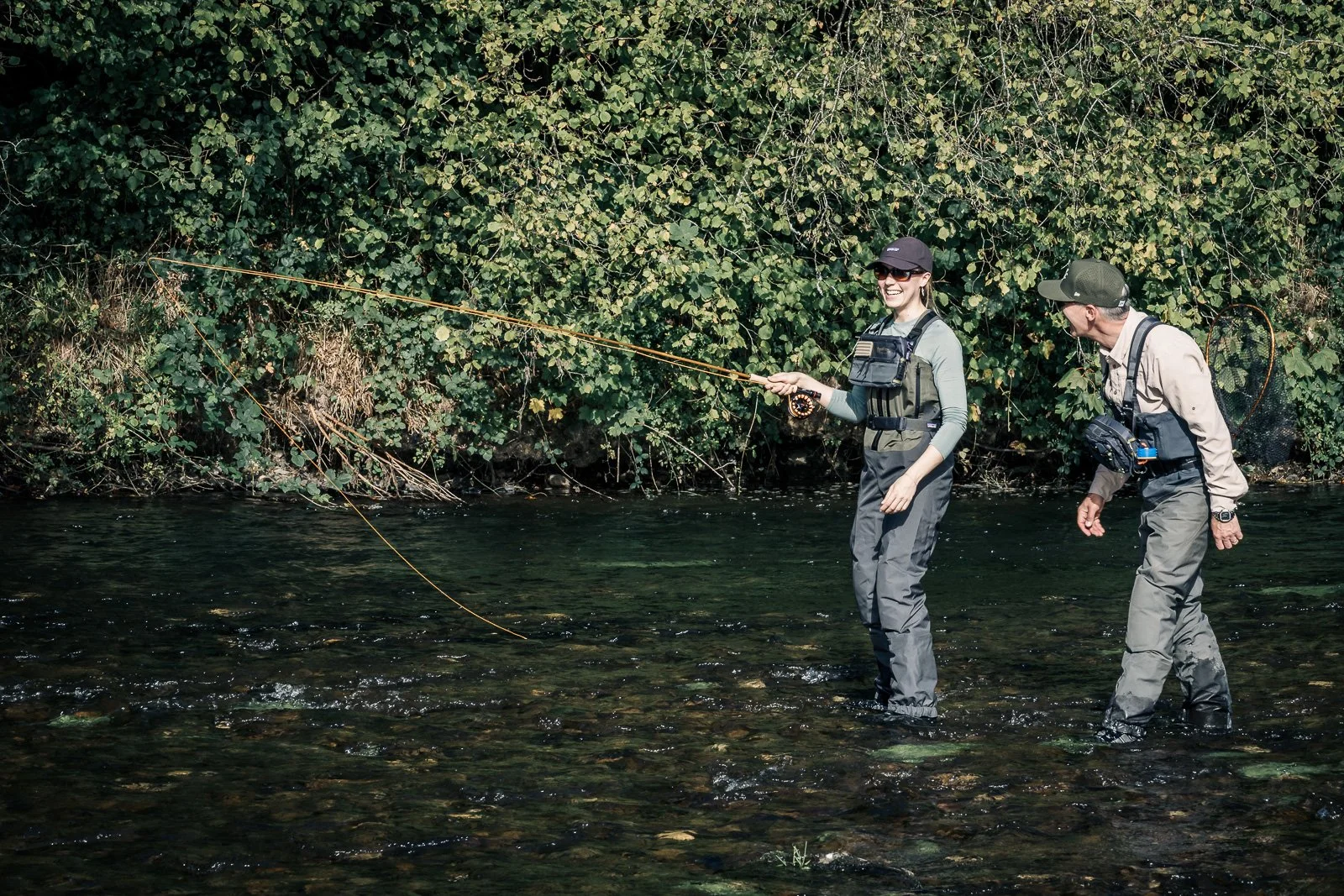 Two anglers fly-fishing in a shallow, clear river surrounded by dense greenery