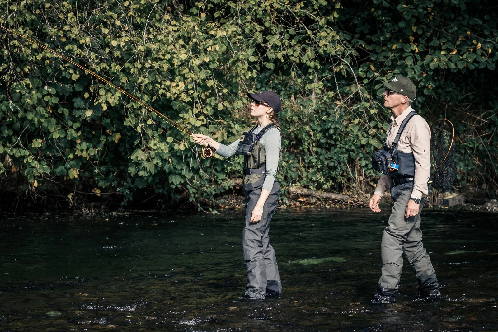 Two anglers fly fishing in waders in a shallow, forest-lined stream