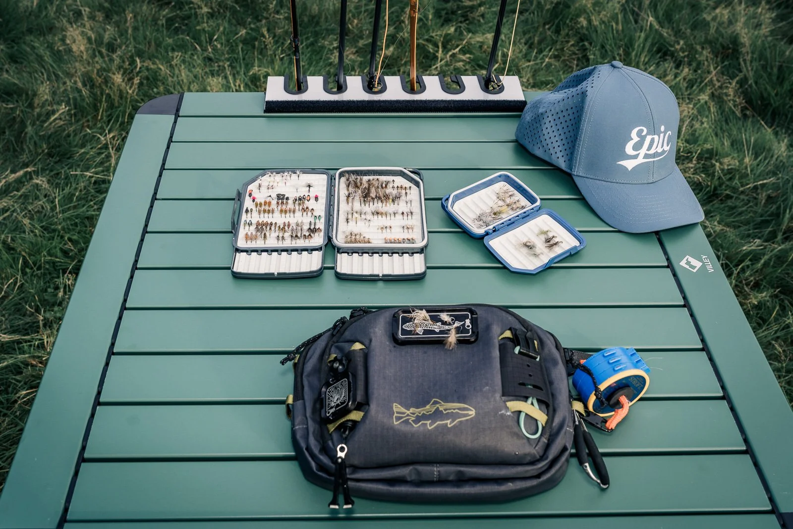 Fly-fishing gear with fly boxes, fishing flies, rods, tackle backpack, and an Epic cap on a green outdoor table