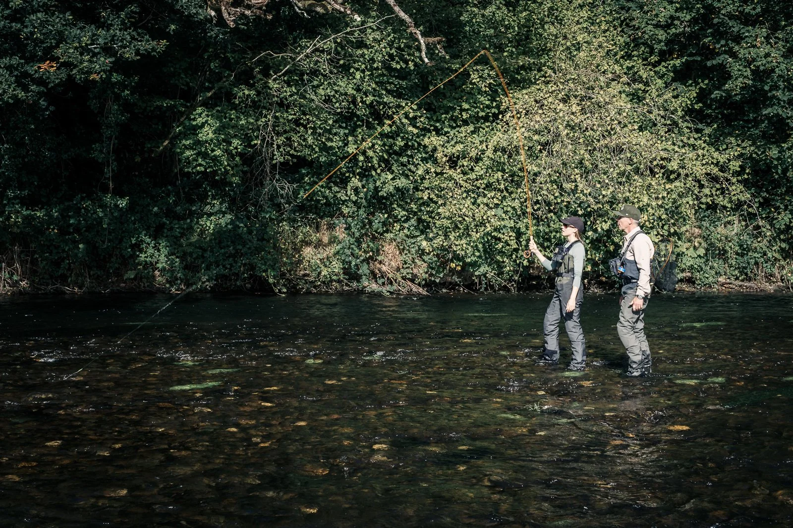 Two anglers fly fishing in waders in a forested river