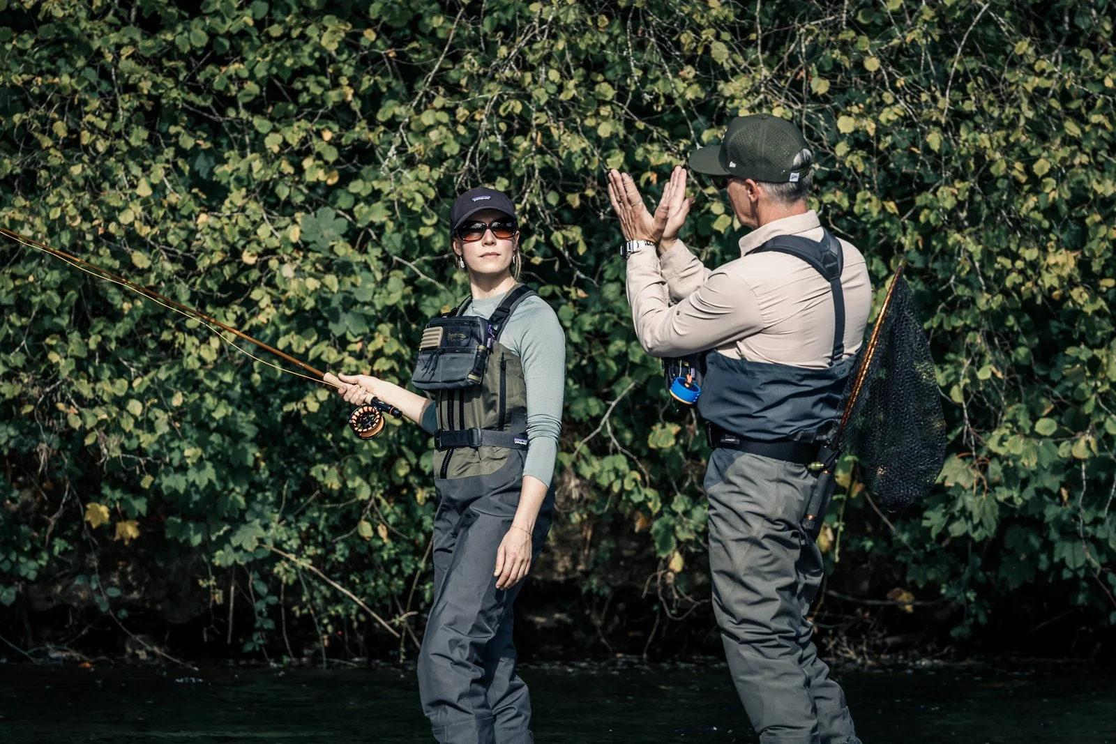 Two fly fishers wading in a river, with one holding a fly rod and the other carrying a landing net.