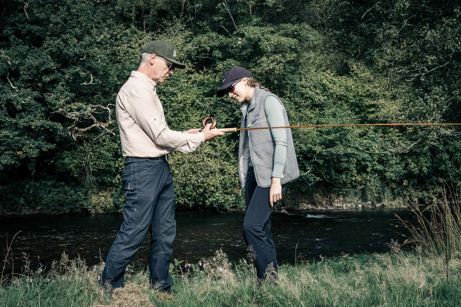 Two anglers beside a forested river, examining a fly-fishing rod and reel.