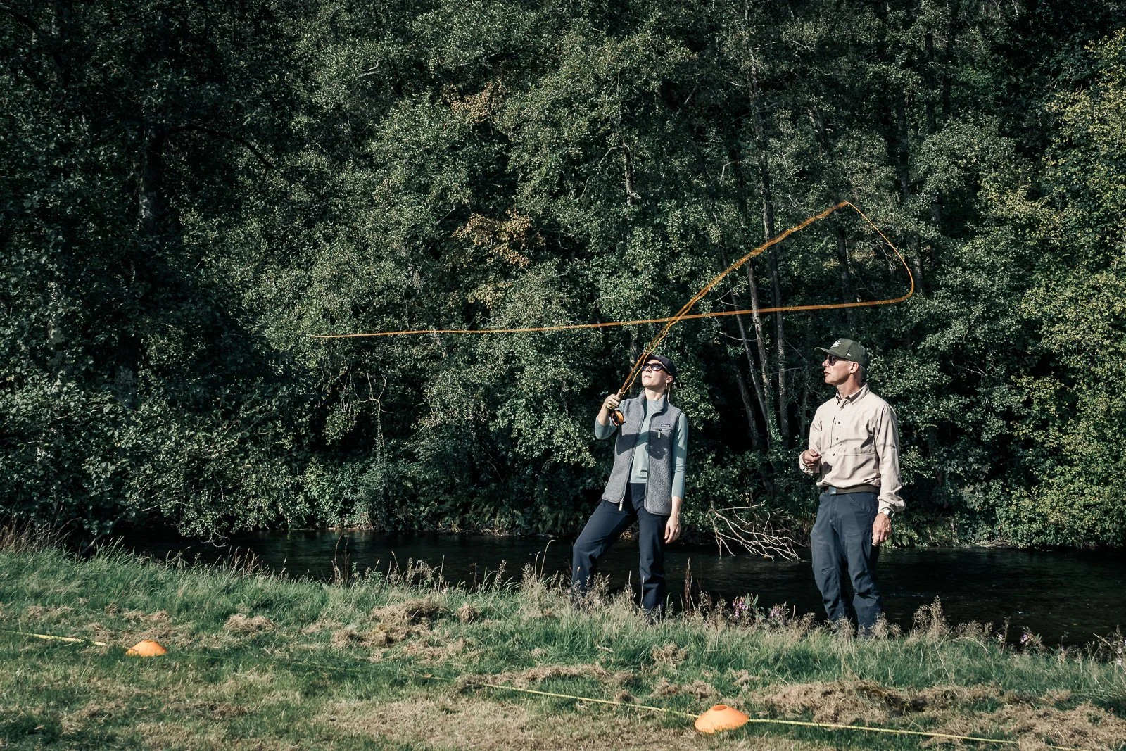 Two people fly-fishing beside a wooded river, with an orange fly line arcing through the air.