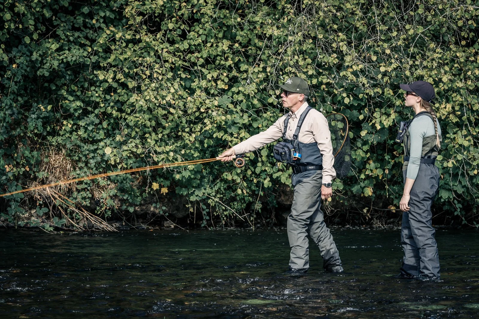 Two fly fishers wading in a shallow river, casting beside a leafy forest bank