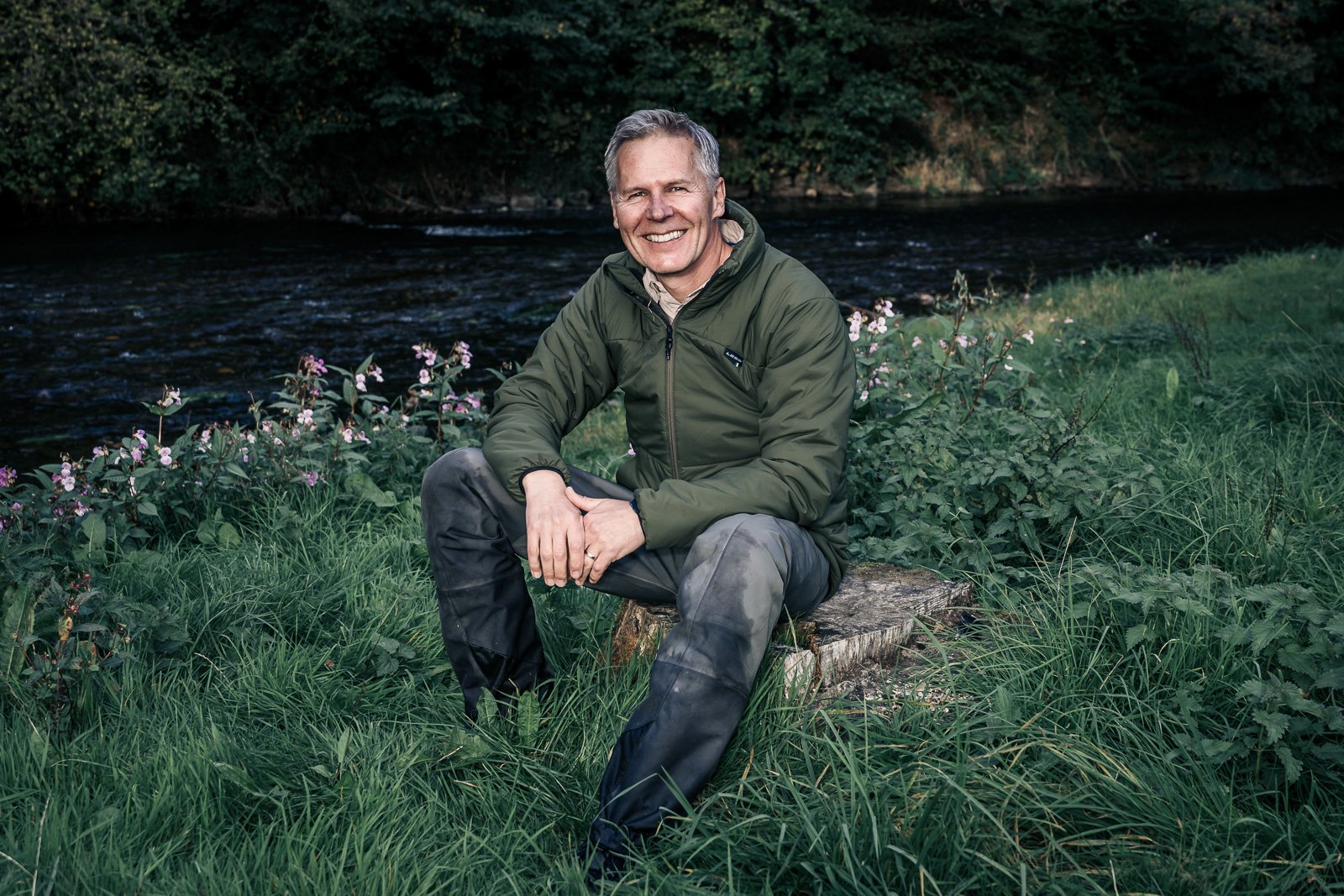 Smiling man in an outdoor jacket sitting on a tree stump beside a river in a grassy woodland setting