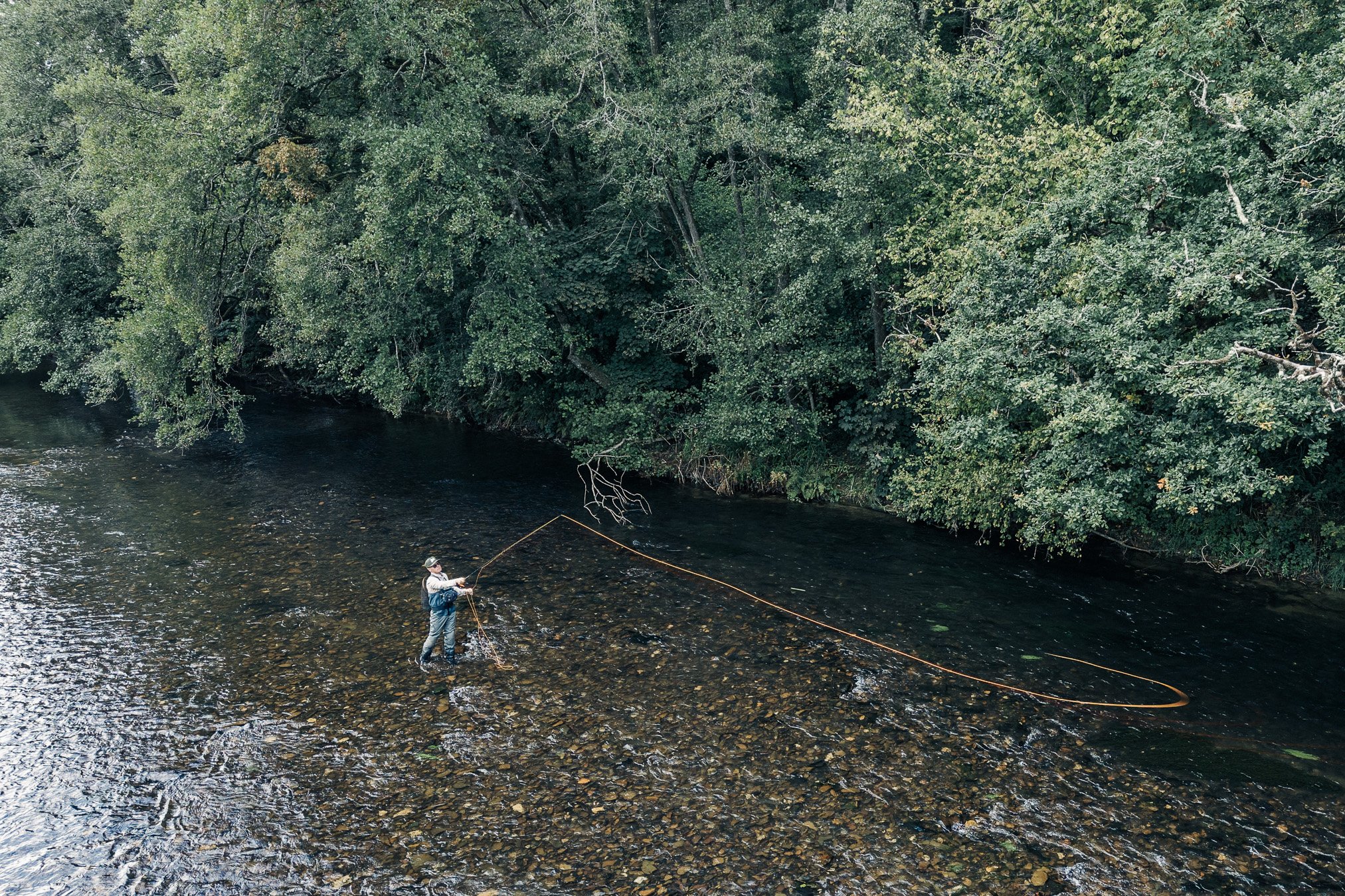 Fly fisherman casting a long line in a shallow, tree-lined river