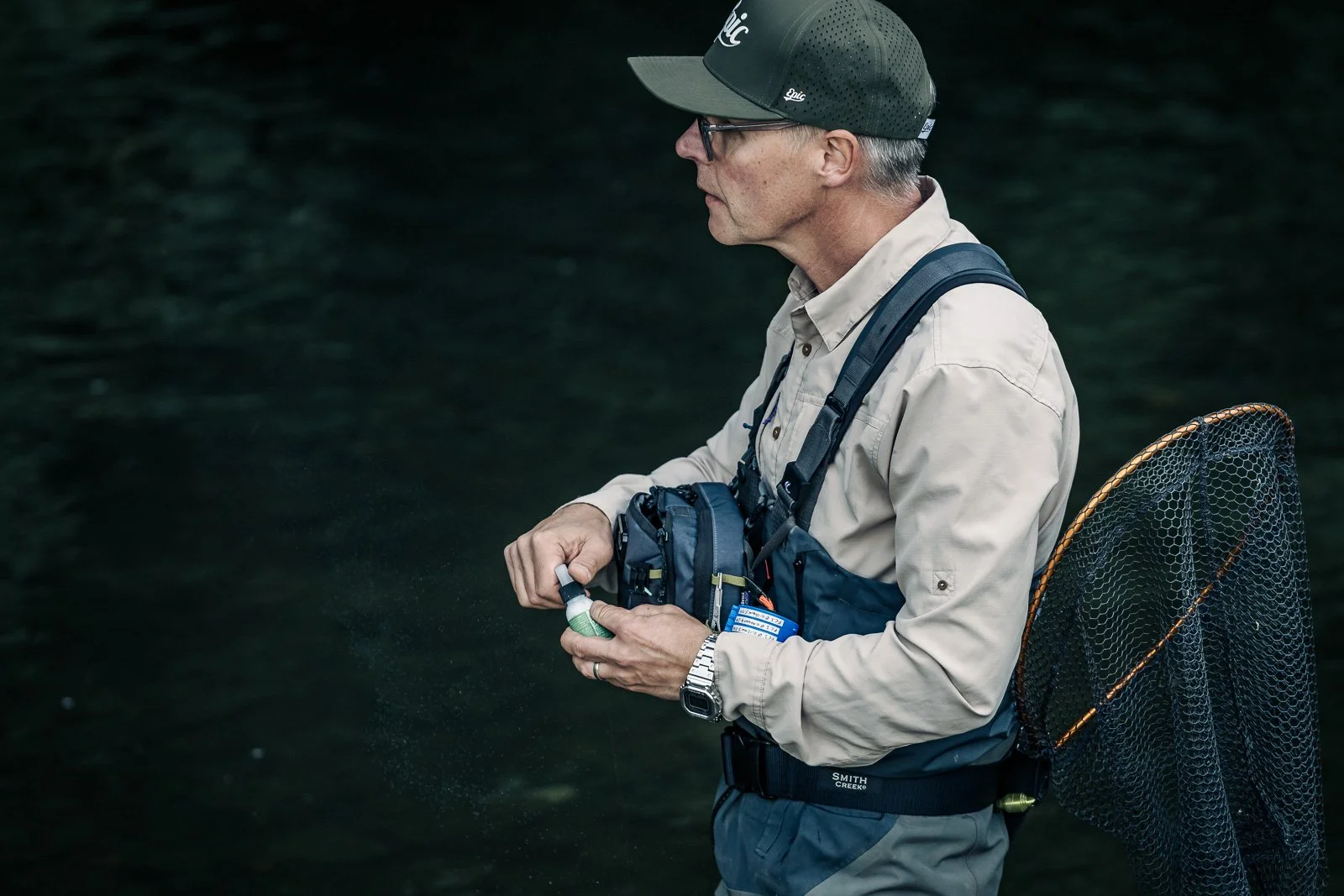 Fly fisherman wearing waders and a cap, holding fishing gear with a landing net beside a dark river