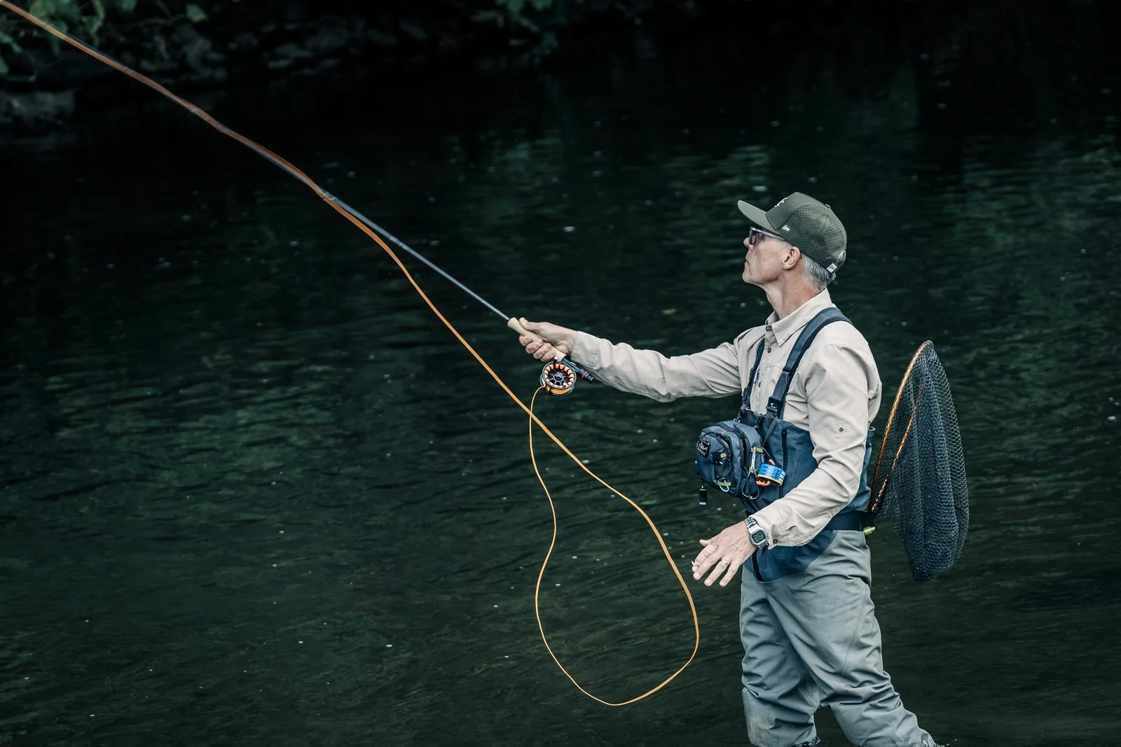 Fly fisherman casting a fly rod in a dark river while wearing waders and carrying a fishing net
