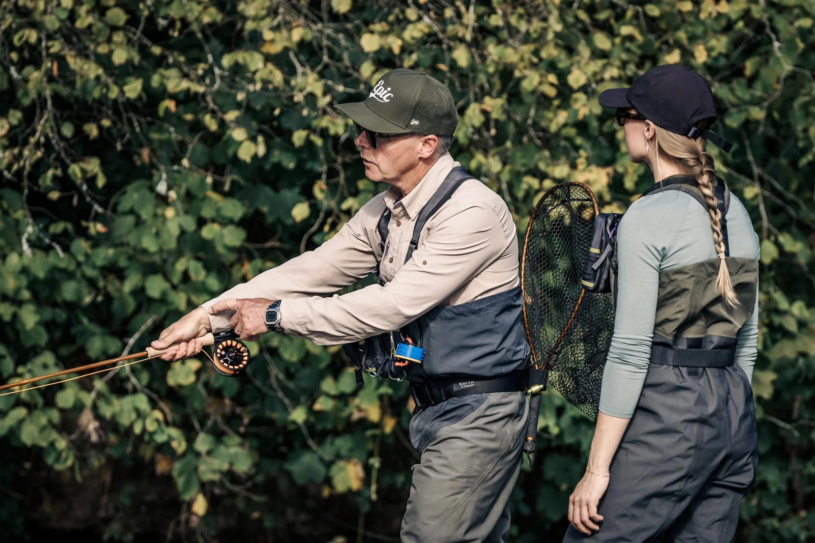 Two fly fishers wearing waders and caps prepare to cast beside a wooded stream, with a landing net visible.