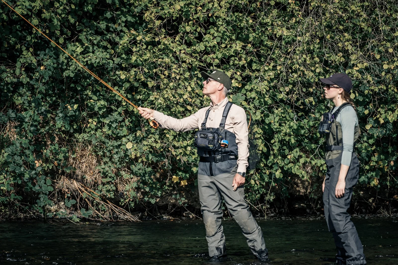 Two anglers fly-fishing in a shallow river surrounded by dense greenery