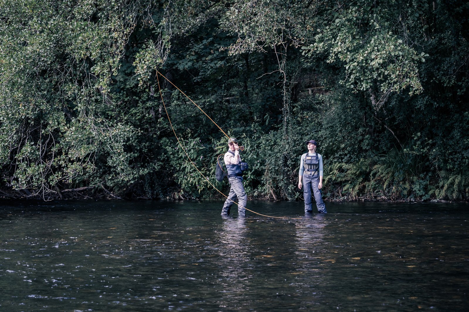 Two fly fishermen wading in a forested river, casting a fishing line.